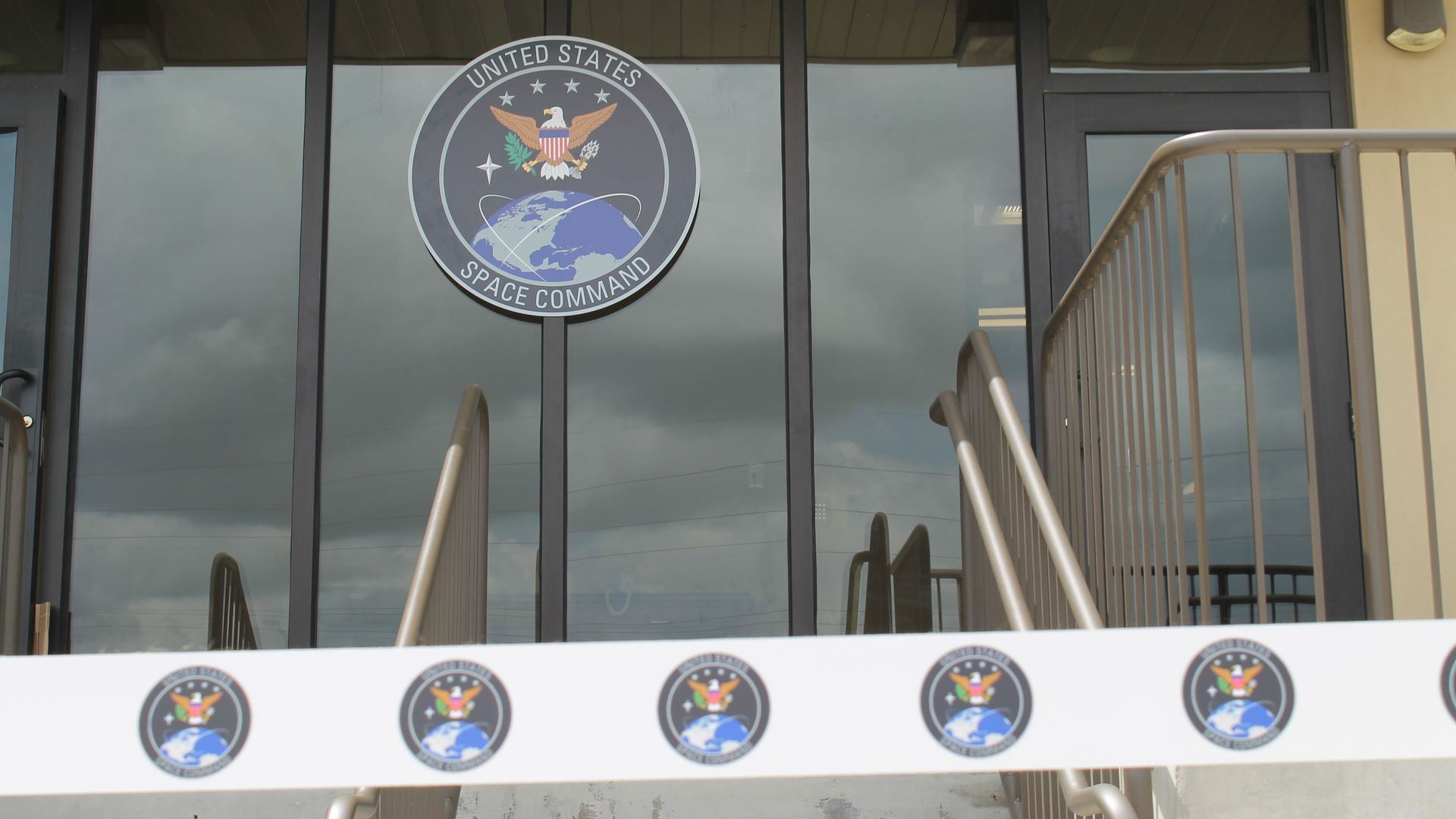 Front view of glass doors with a large circular United States Space Command emblem featuring an eagle, stars, and a globe; metal railing and beige wall nearby; reflections in the glass.