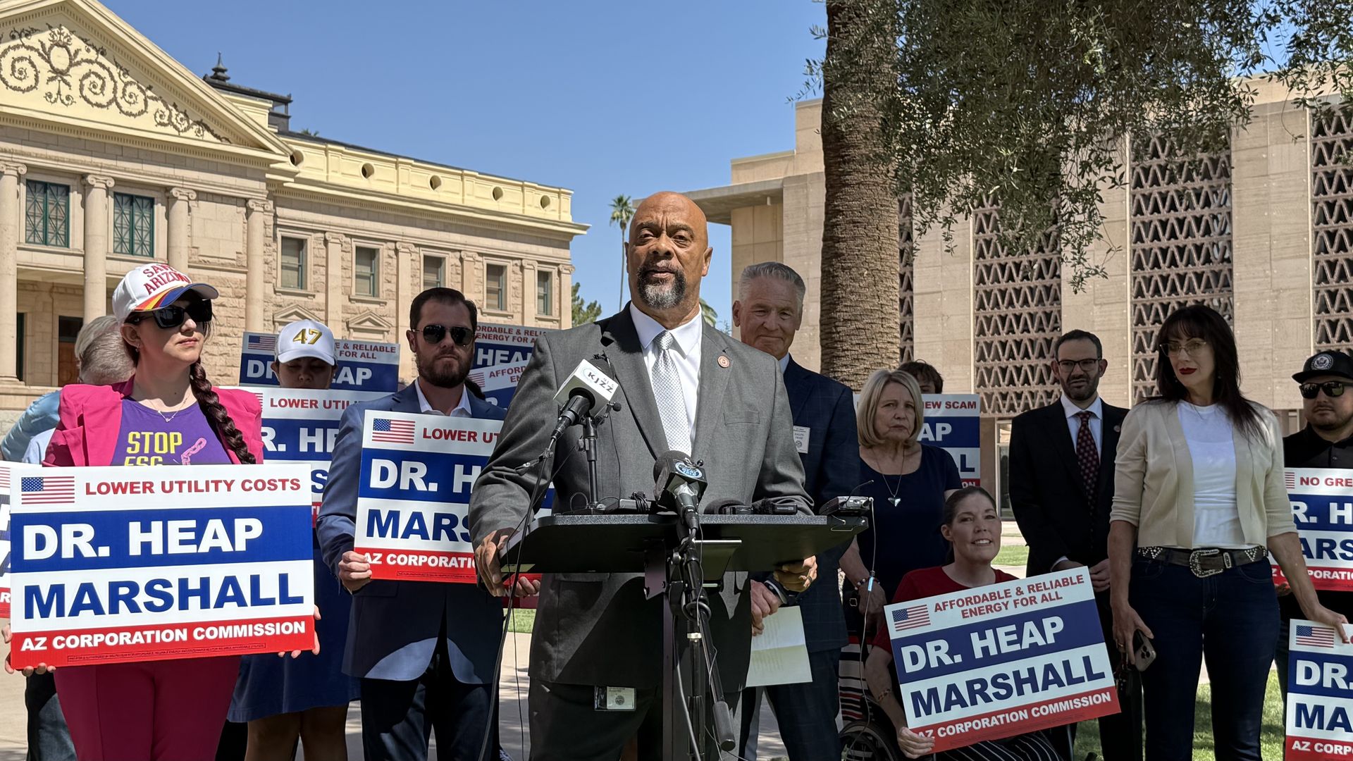 Outdoor press conference with a bald man in a gray suit at a podium, flanked by supporters holding blue and white signs reading "DR. HEAP MARSHALL" for the Arizona Corporation Commission; blue sky and the Arizona Capitol are in the background.