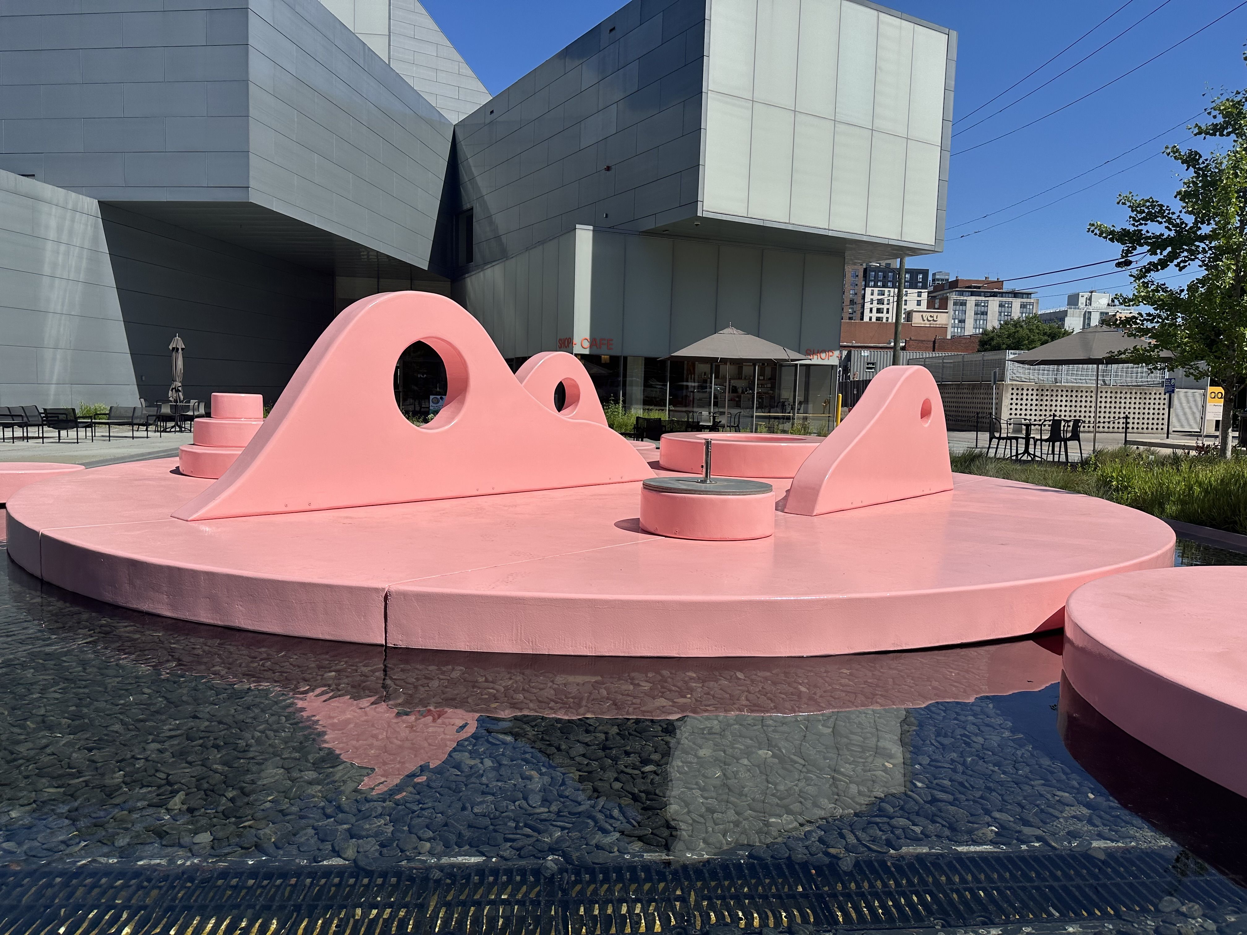 Outdoor scene of a modern gray angular building behind a large pink sculpture and circular pink fountain. Clear blue sky, trees, outdoor seating, and water in a shallow pool.