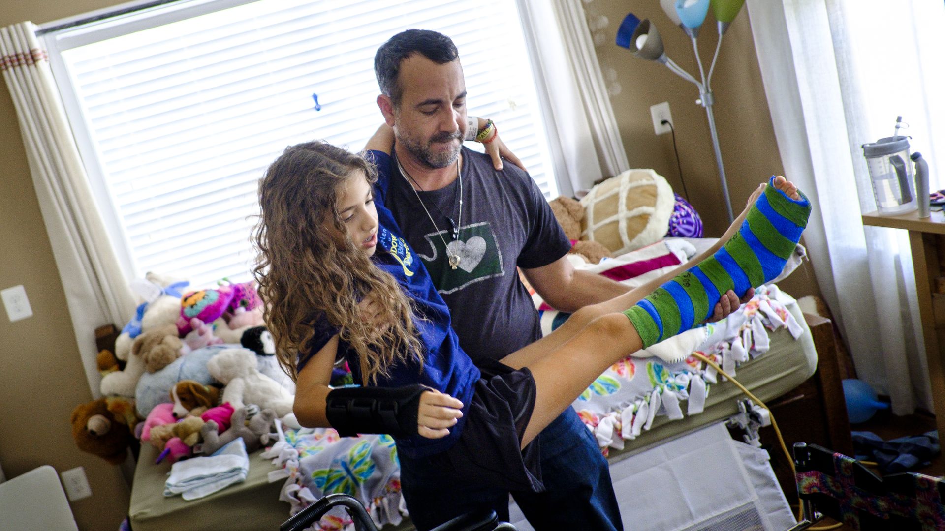 A father carries his daughter, who has a cast, in her hospital room.