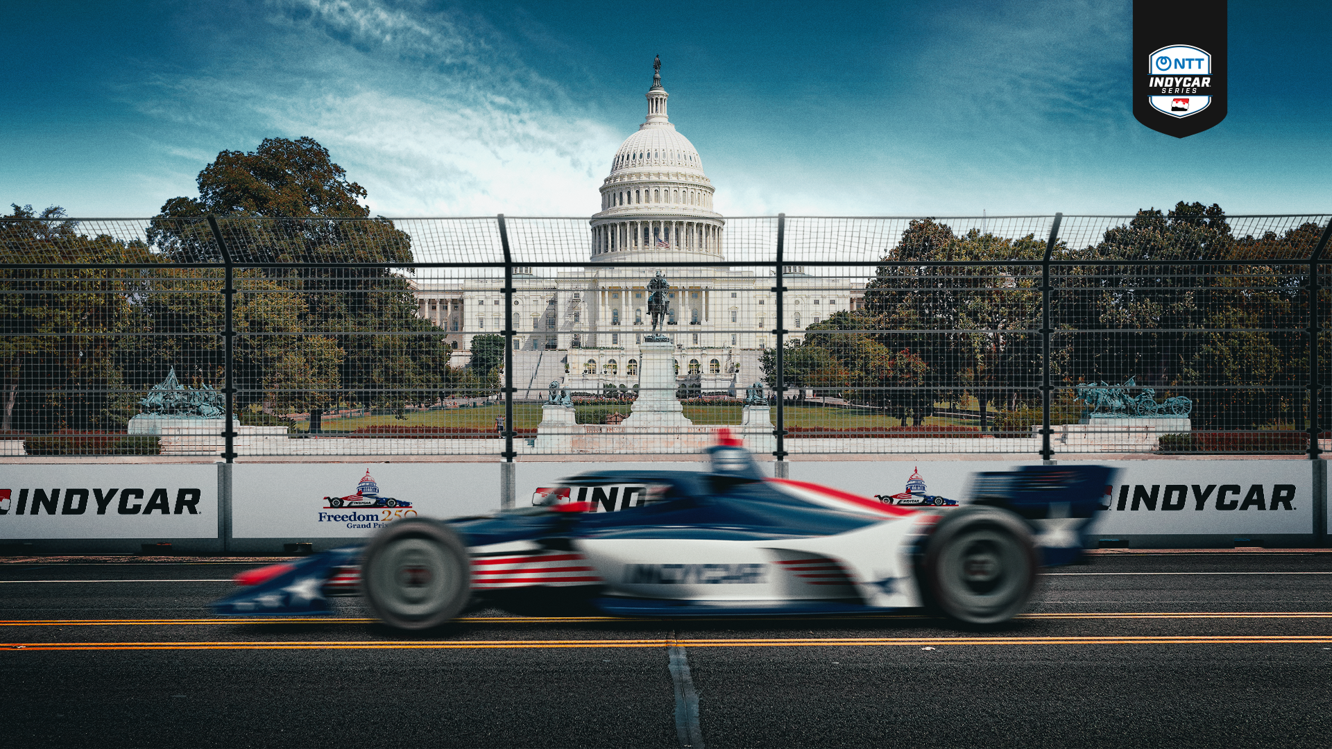 Blurred IndyCar racing on a track with safety fences, U.S. Capitol dome and statue in background, under a blue sky. Signs show IndyCar and Freedom 250 Grand Prix logos.