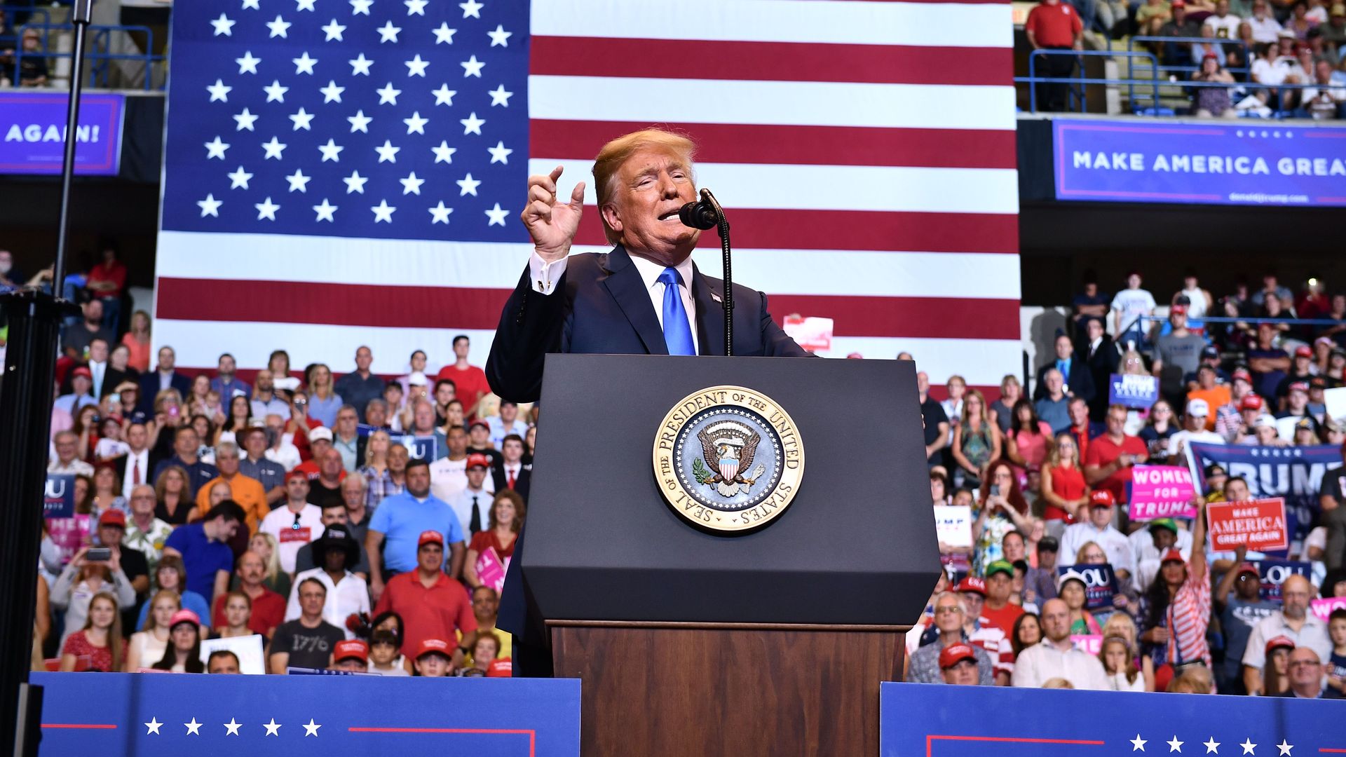 Donald Trump speaks from a podium, in front of a crowd and large American flag