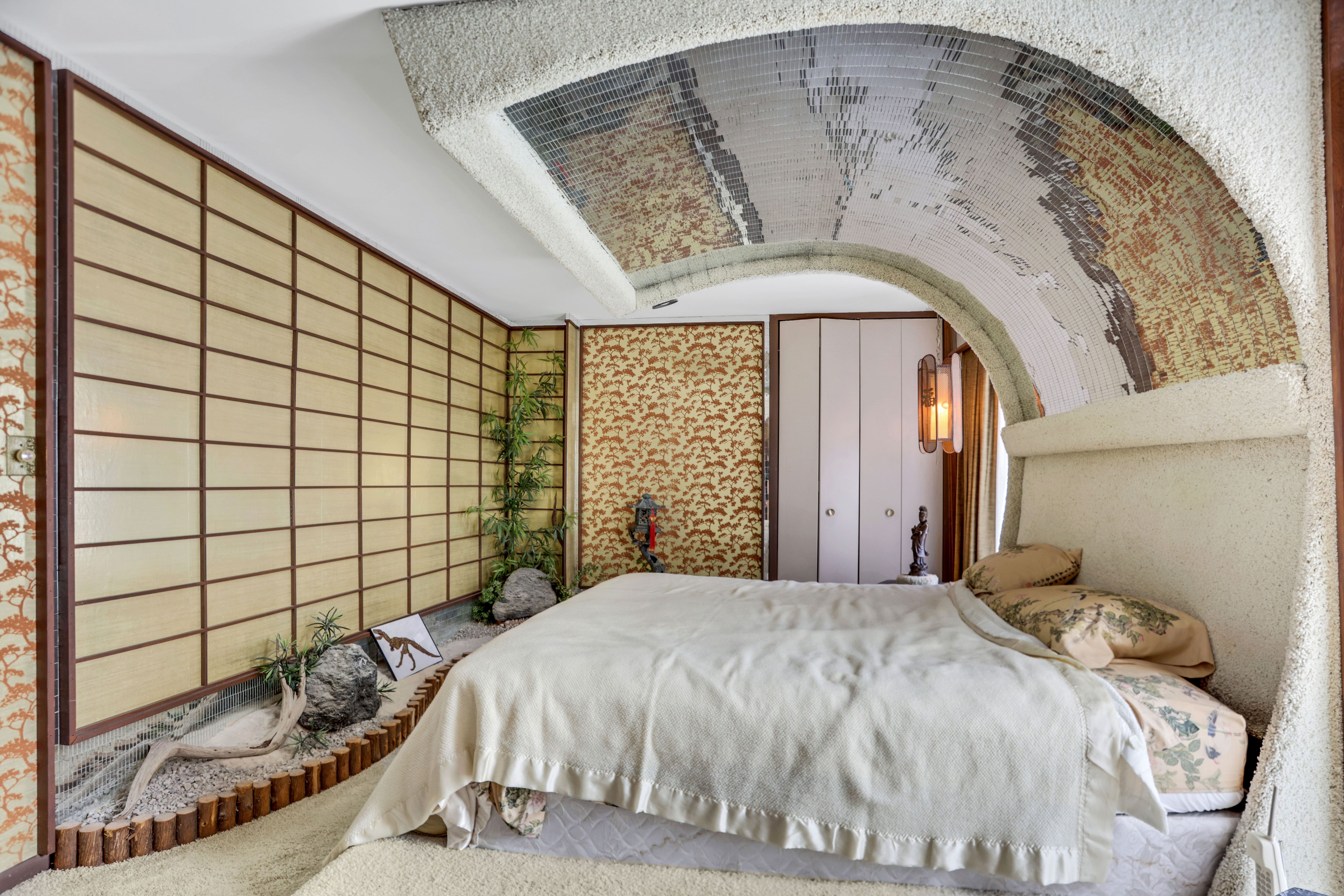 A bed with a wave-like headboard covered in mirrored tiles that stretches to the ceiling, facing an interior rock garden and Japanese walls