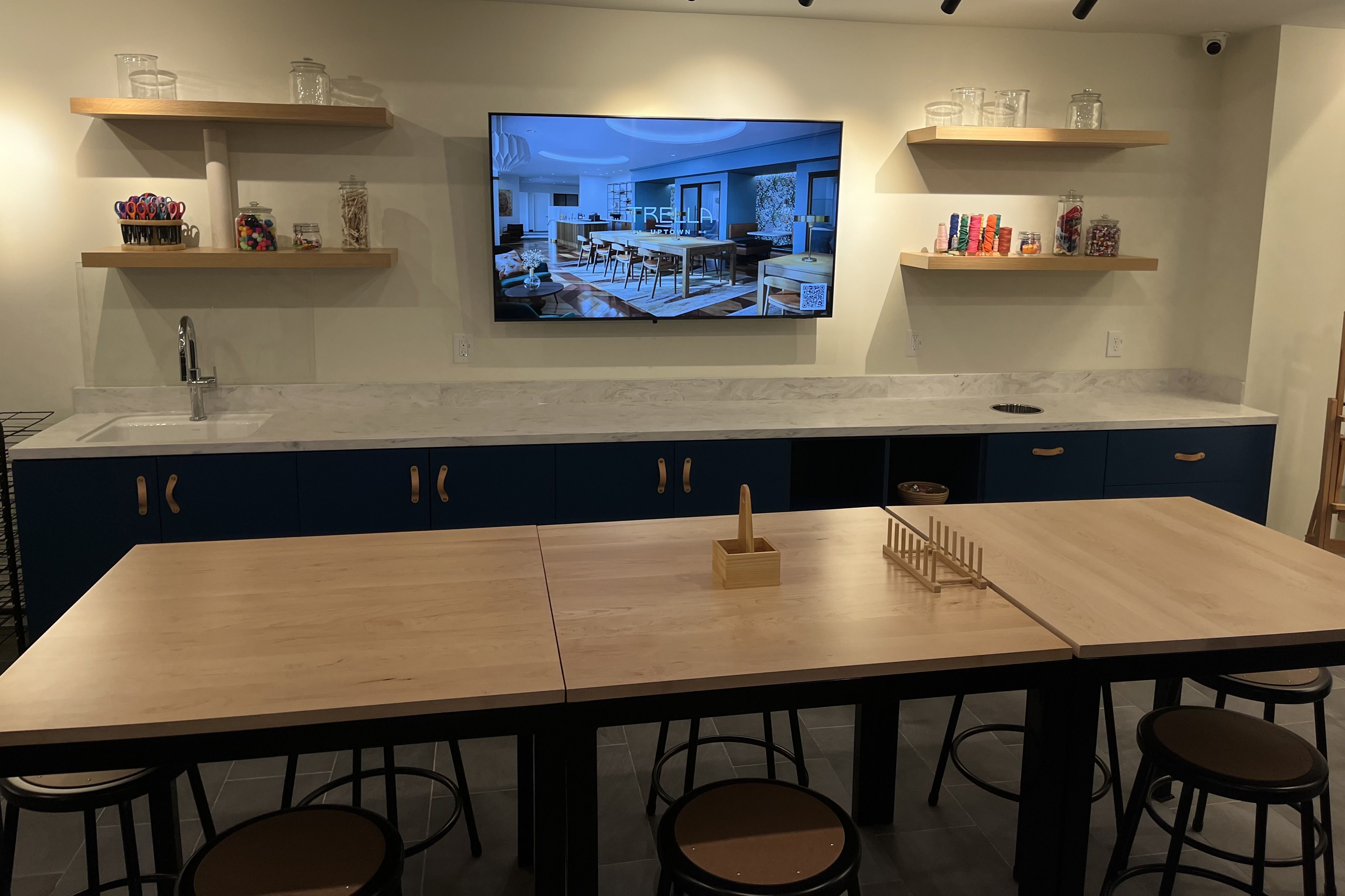 Craft room with blue lower cabinets, light wooden tables, stools, floating shelves with jars of craft supplies, and a TV showing a kitchen design with "TRELLA UPTOWN".