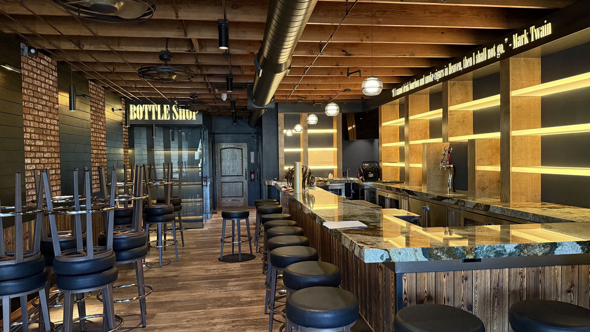 Empty bar interior with wooden ceiling and floors, polished marble bar, black cushioned stools, illuminated shelves, and a sign reading "BOTTLE Shop" and a Mark Twain quote lit above the bar.