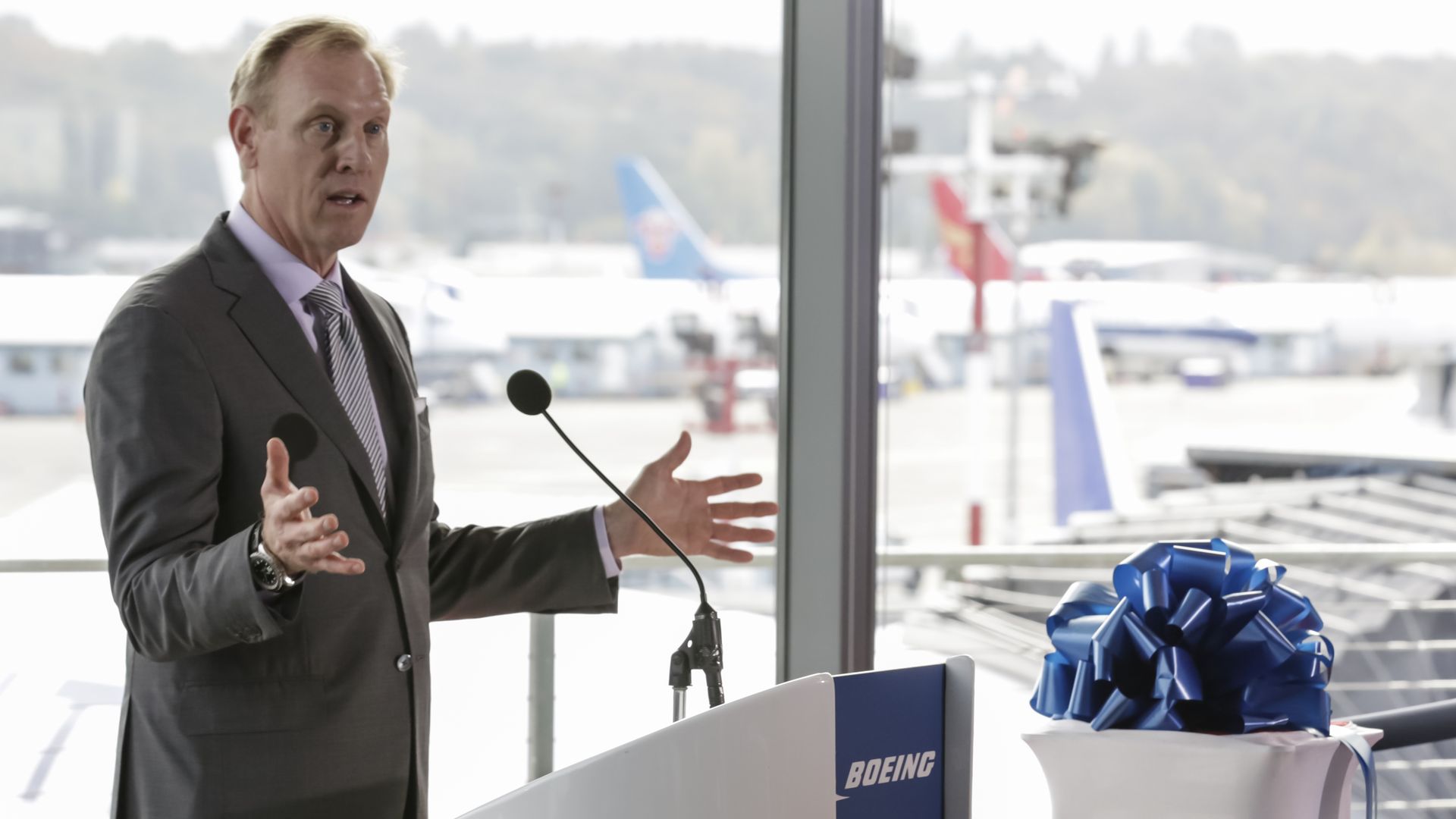 In this image, Pat Shanahan speaks in front of a microphone and podium. The windows behind him look out onto an airport.