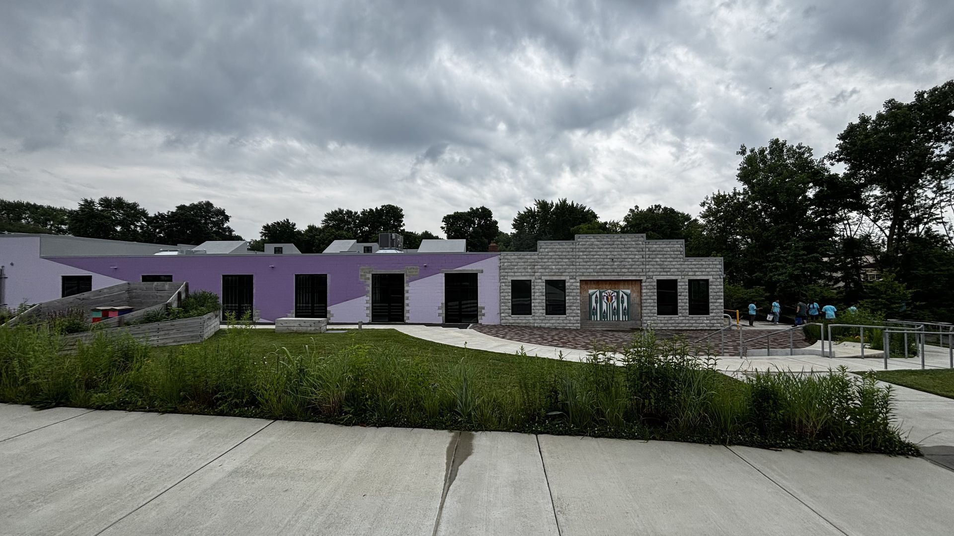 Wide view of a modern building with purple and gray walls, black windows, and a decorative door, surrounded by green grass and plants under a cloudy sky, with people in blue shirts walking away.