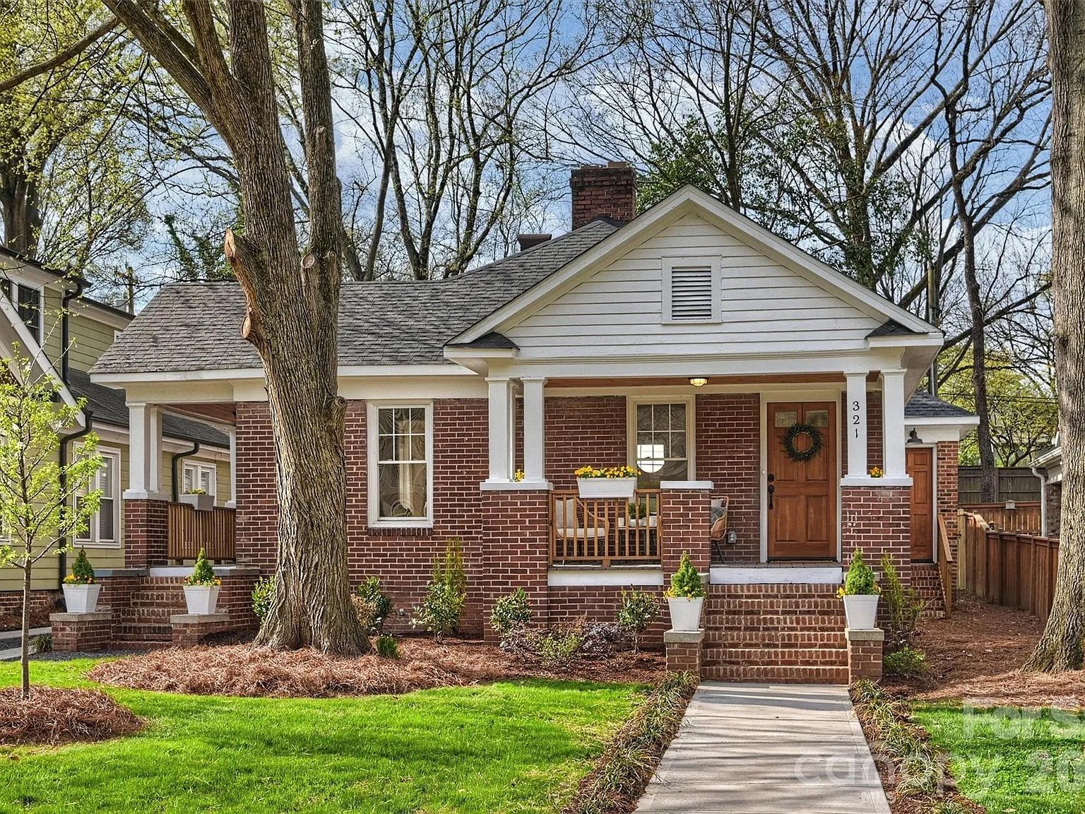 Charming single-story brick cottage with white trim, a covered front porch, brick steps, a wreath on the door, and a green lawn with tall trees and planters.
