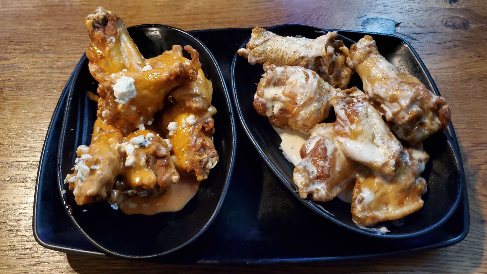 A black rectangle plate with two bowls of chicken wings on it, in sauce