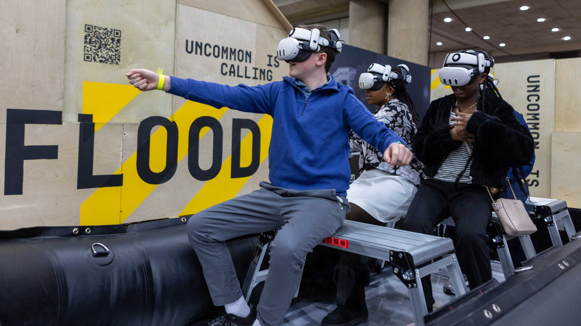 Three people wearing VR headsets ride an inflatable raft in a tech exhibit. A man in a blue hoodie extends his arms forward while two others watch; background shows yellow diagonal stripes and the words "UNCOMMON IS CALLING".