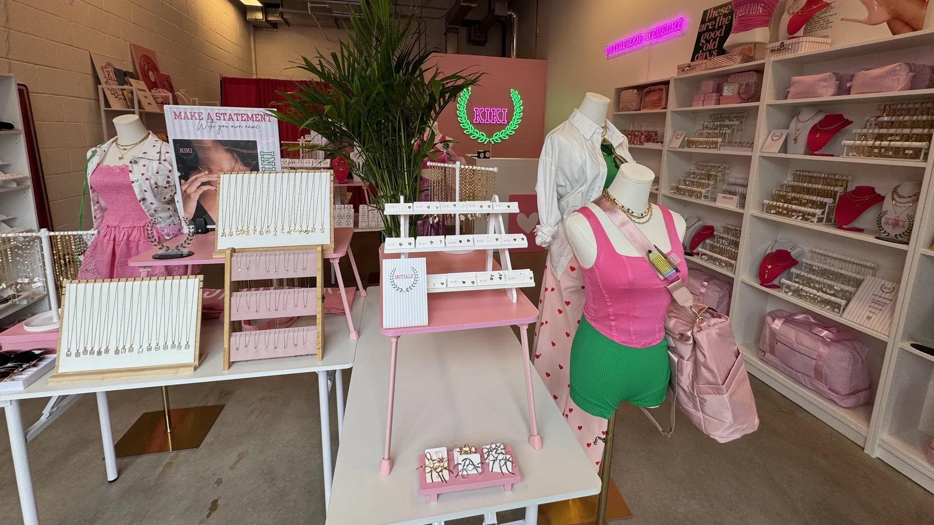 Pink-themed jewelry shop interior with necklace displays, mannequins in pink tops and green shorts, a tall plant centerpiece, shelves of accessories, and a neon "KIKI" sign.