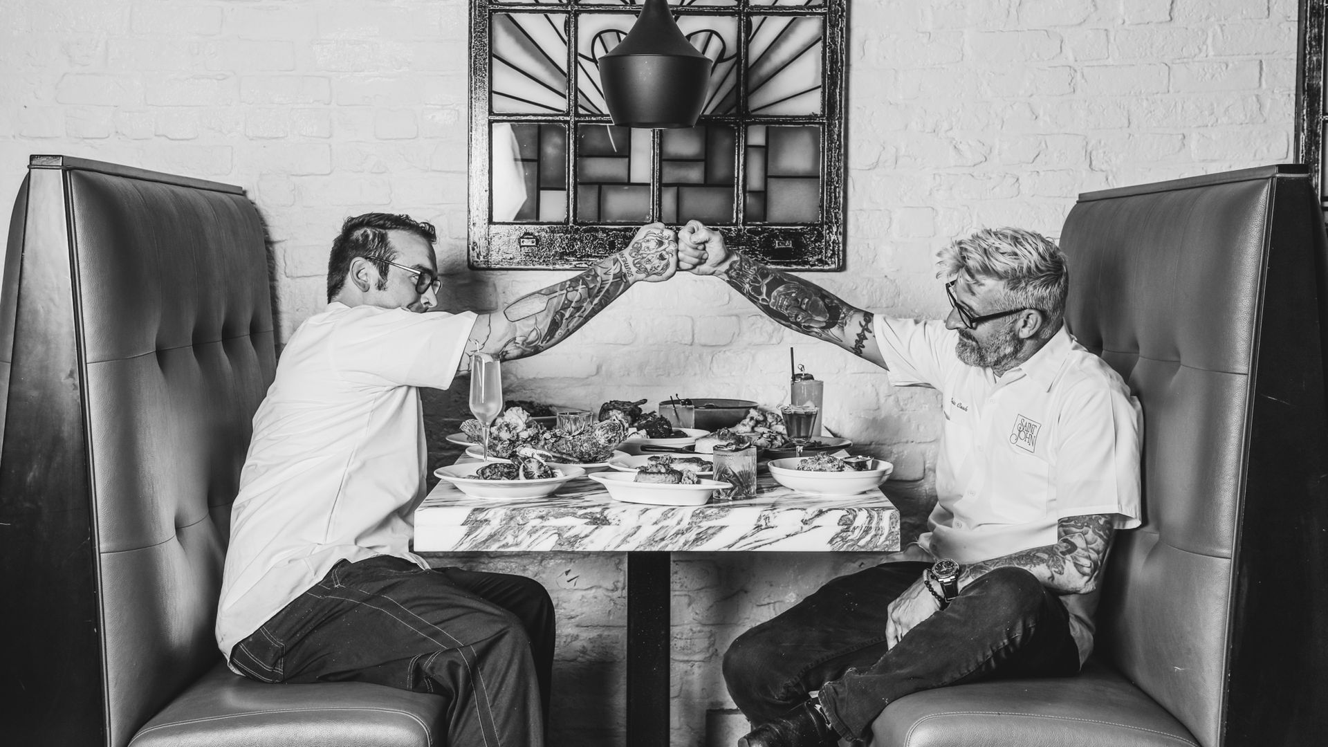A black and white image of Saint John chef de cuisine Daren Porretto and executive chef Eric Cook fist-bumping across a table while sitting in a booth at Saint John. The table is filled with plated food.