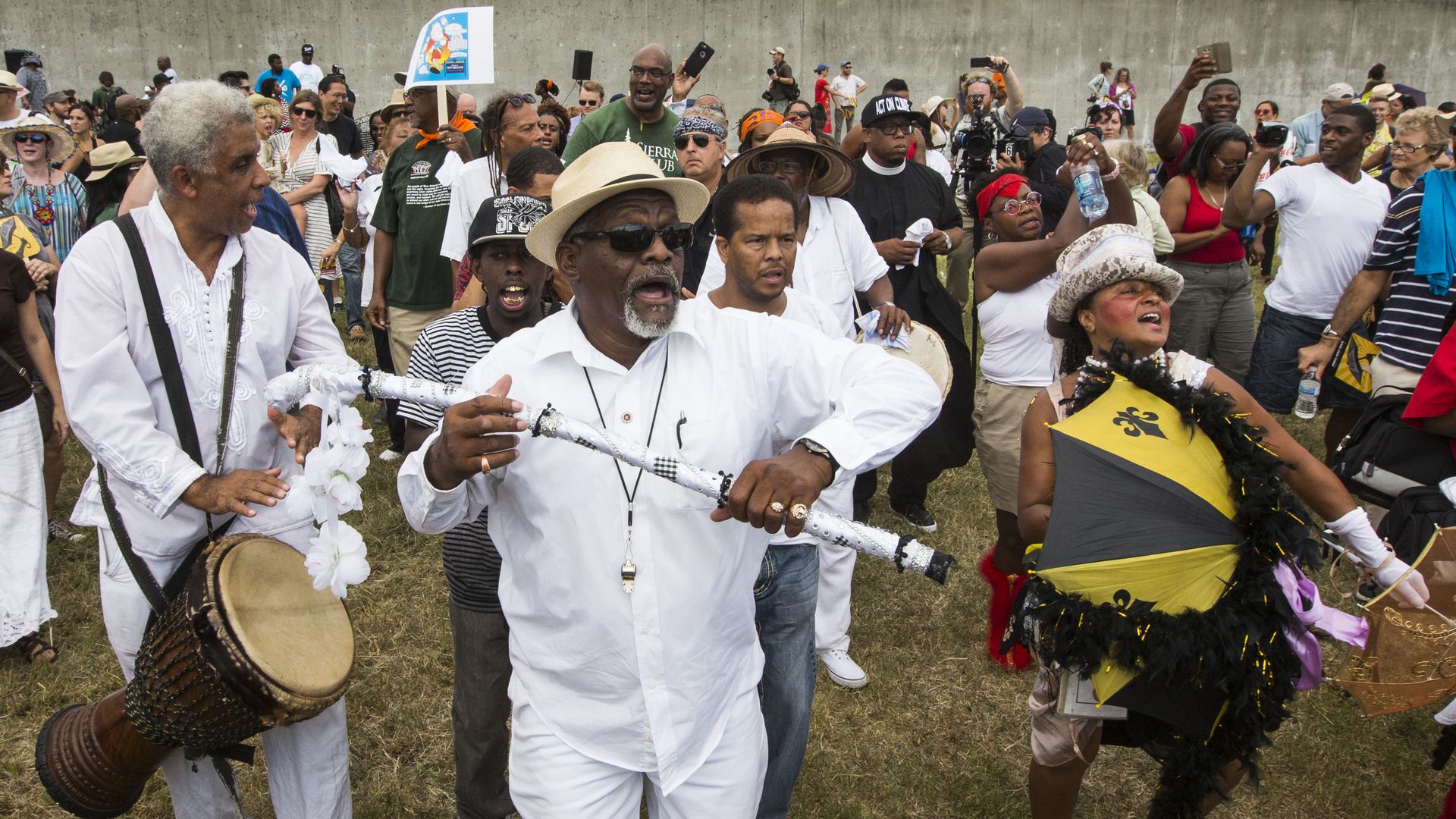 A lively crowd outdoors, featuring a man in white playing a drum, another man in white with a hat and sunglasses holding a decorated cane, and a woman dancing with a black and yellow umbrella.