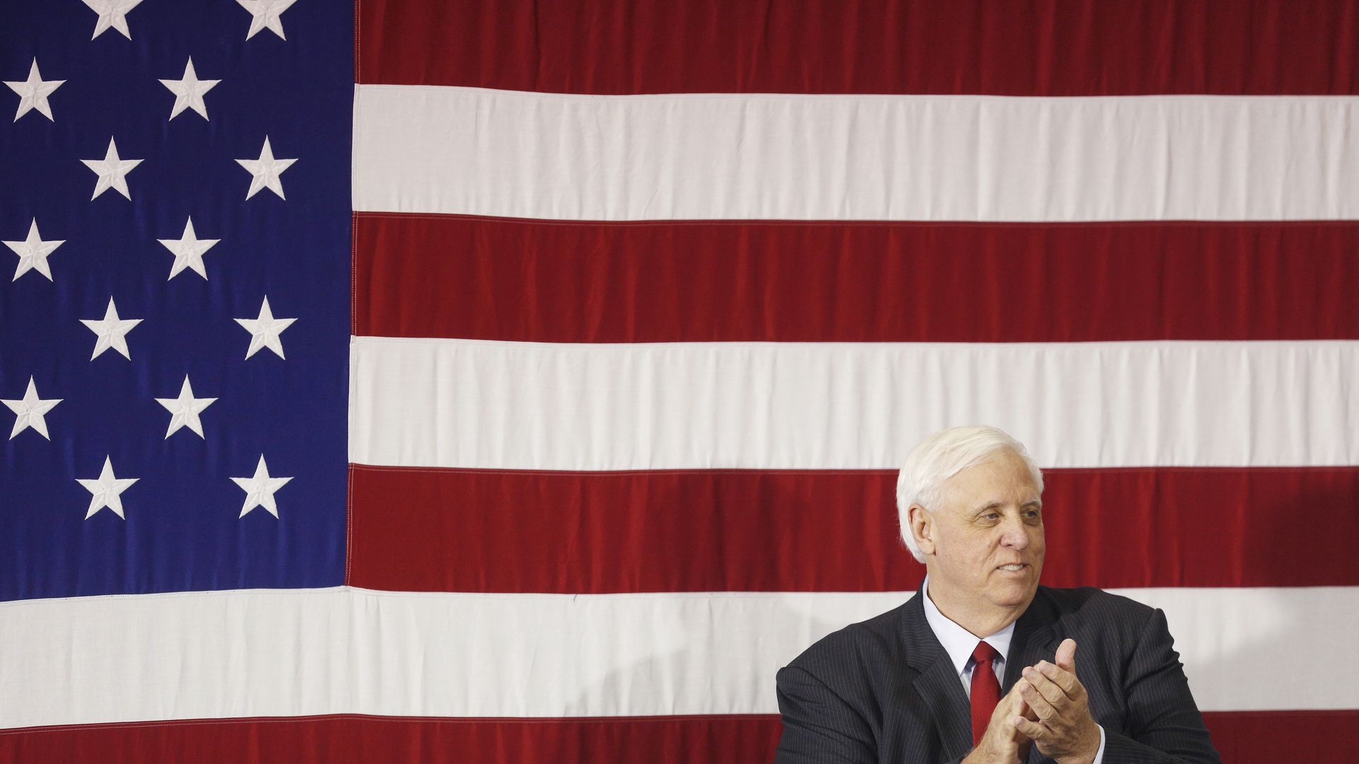 Photo of Jim Justice clapping as he stands against the image of an American flag