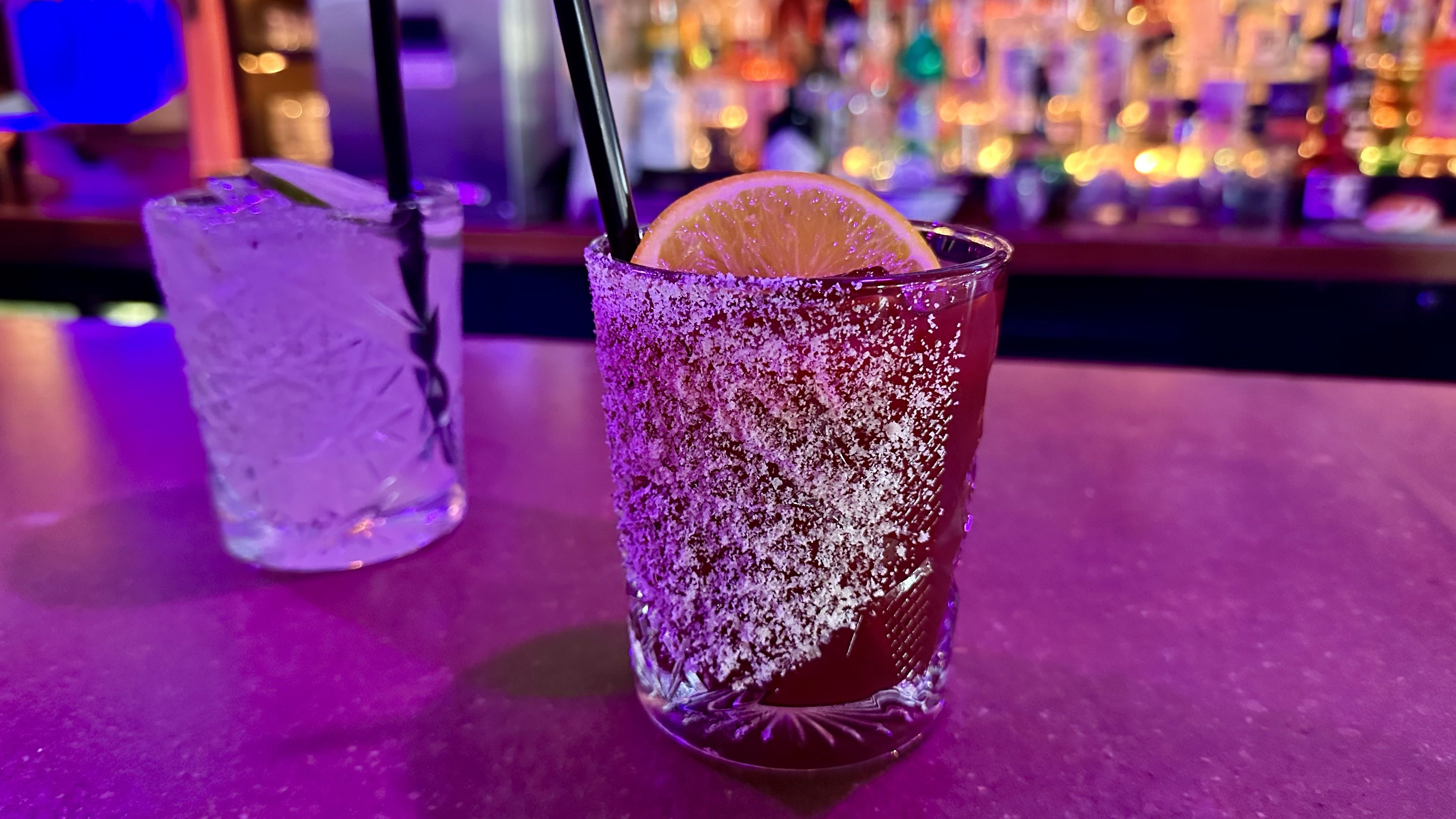 Two cocktails on a neon purple bar: a dark red drink with a salted rim and orange slice in the foreground, and a clear ice-filled glass with a straw to the left; colorful, blurred bar in the background.
