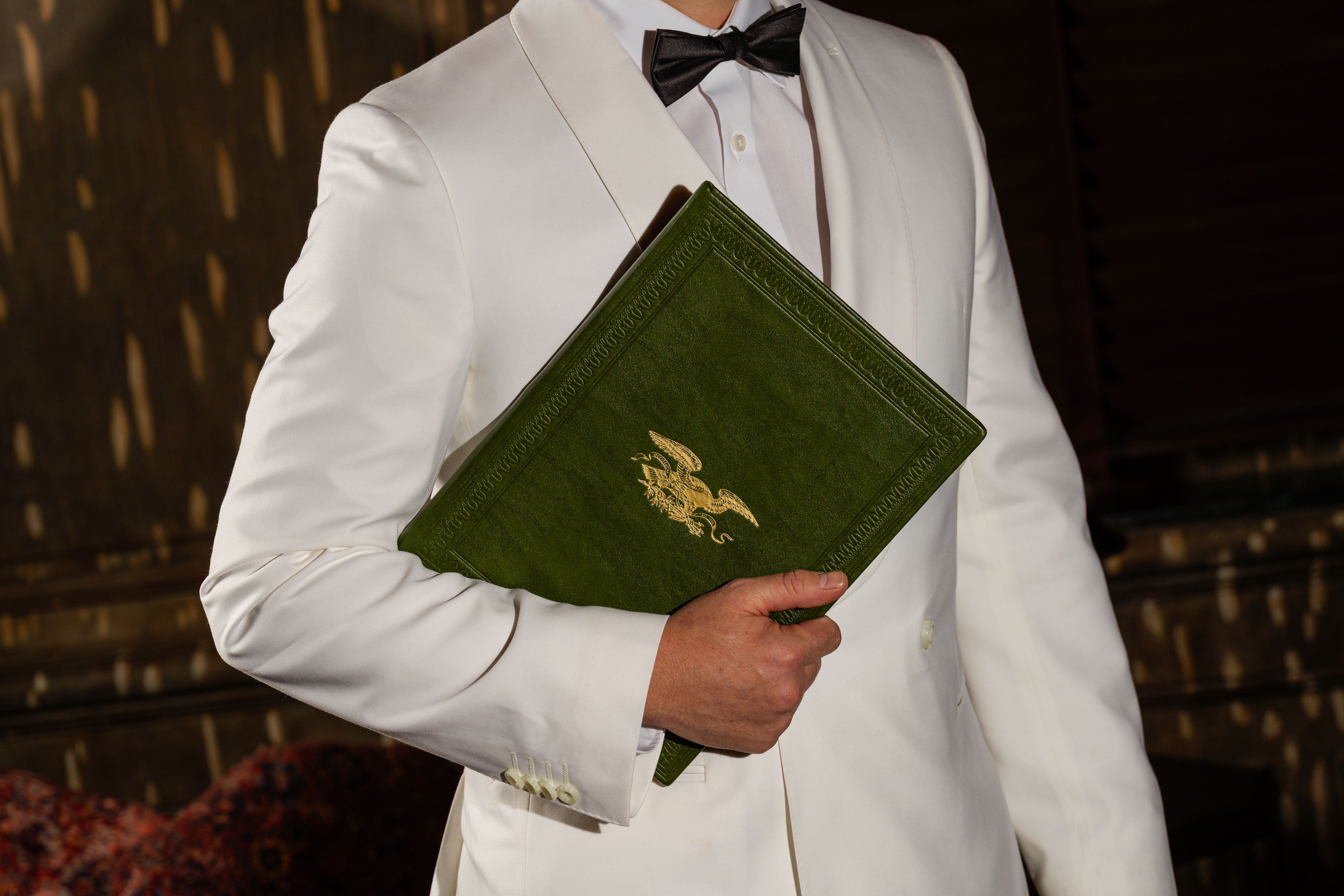 A waiter in a white tuxedo jacket holding a green menu