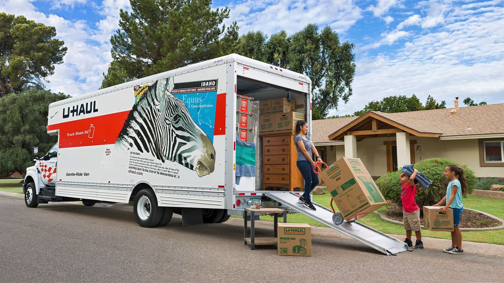 A woman and two children unload a U-Haul truck parked in front of a home. 