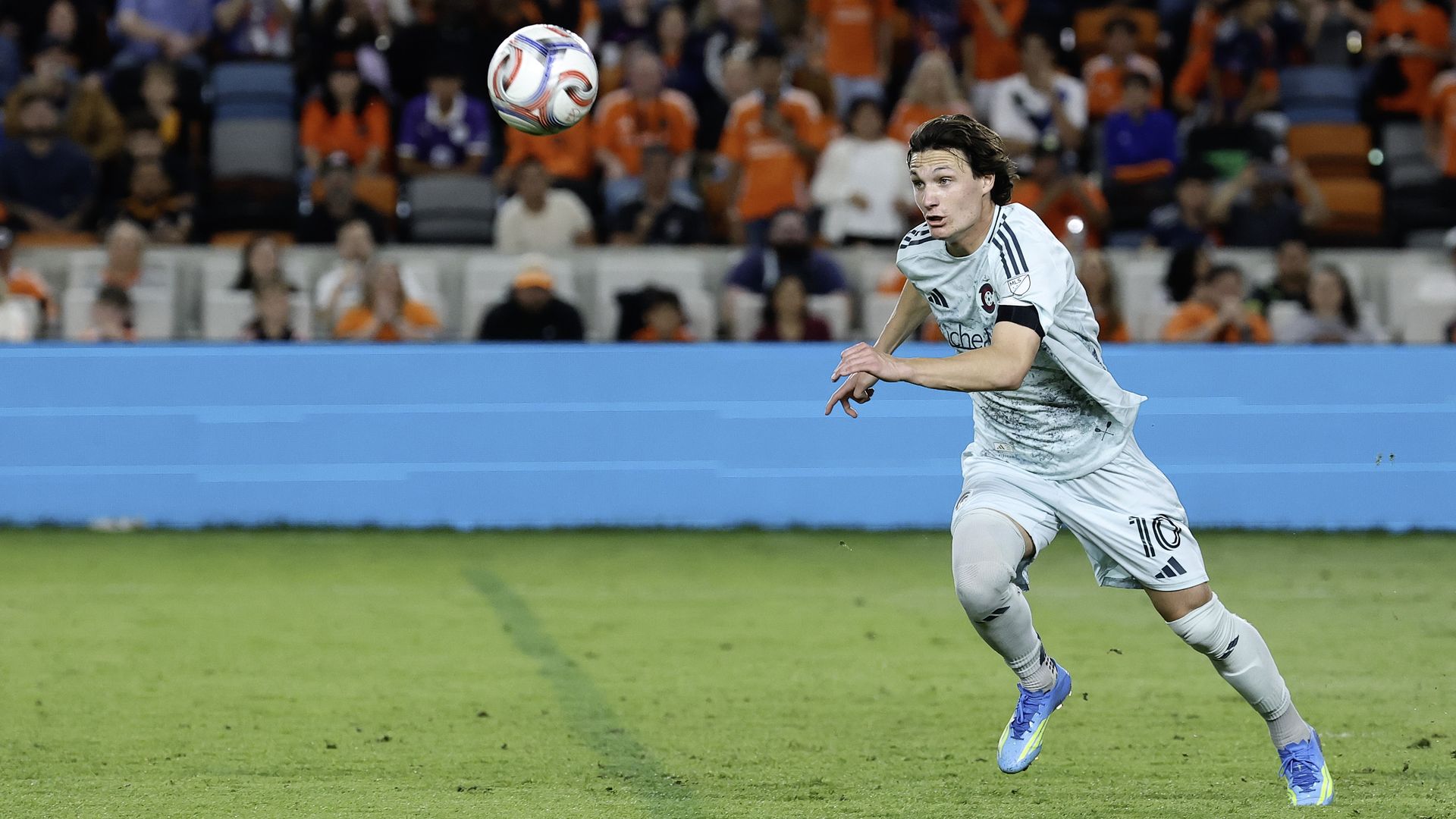Soccer player in a light gray kit with number 10 chases a white ball mid-air on a green pitch; a crowded stadium with orange and blue stands looms in the background.