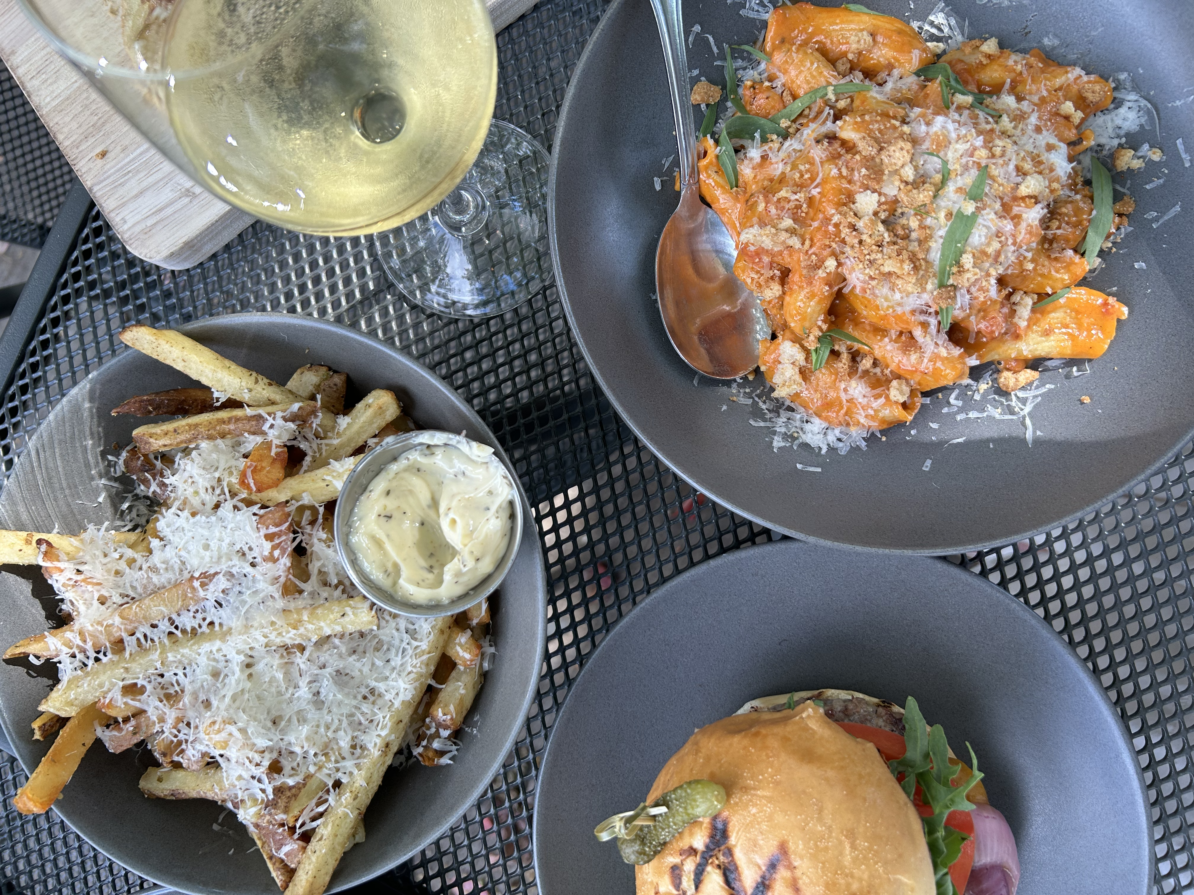 Overhead view of a meal on a black metal table: a glass of white wine, a bowl of fries with grated cheese and dipping sauce, a plate of orange pasta with herbs, and a burger with pickles.