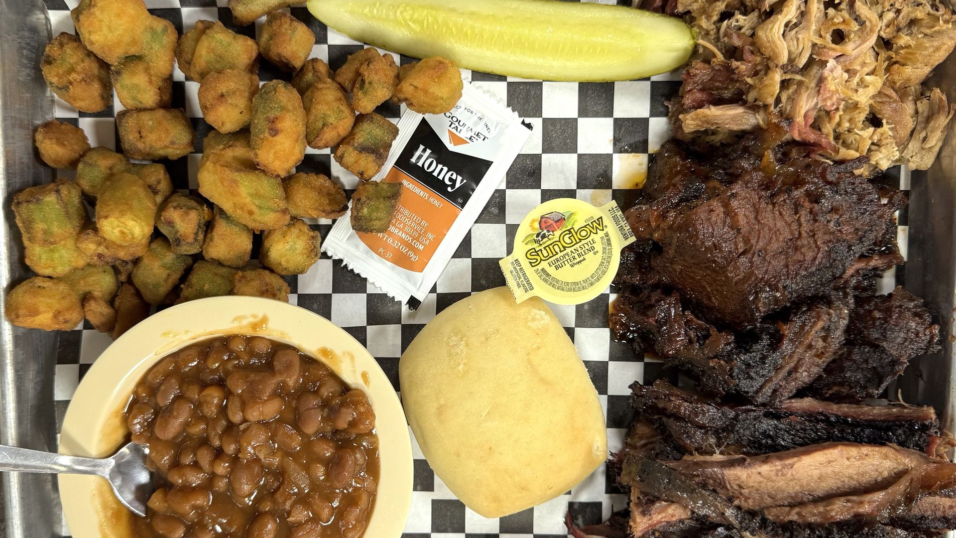 A barbecue platter with smoked brisket, pulled pork, baked beans, fried okra, a dinner roll, pickle spear, honey packet, and butter cup, all served on a checkered paper-lined tray.