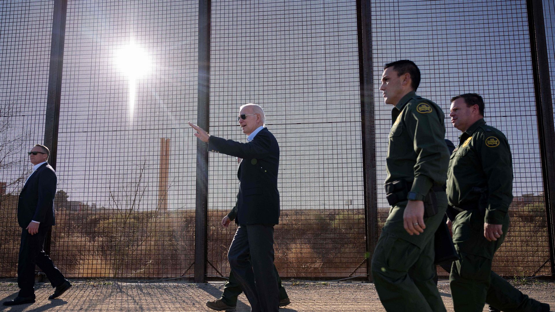 President Biden walks past a tall border barrier, waving with his left hand, while two Border Patrol agents in green uniforms walk behind him.
