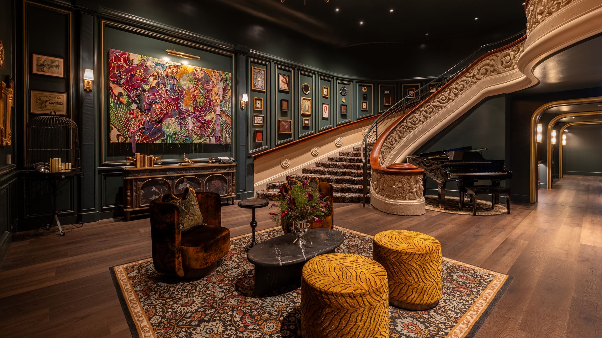 Elegant living room with dark teal walls, an ornate white curved staircase, a grand piano, a large colorful painting with framed art, chandelier lighting, and a seating area of brown velvet chairs, yellow tiger-striped ottomans, and a patterned rug.