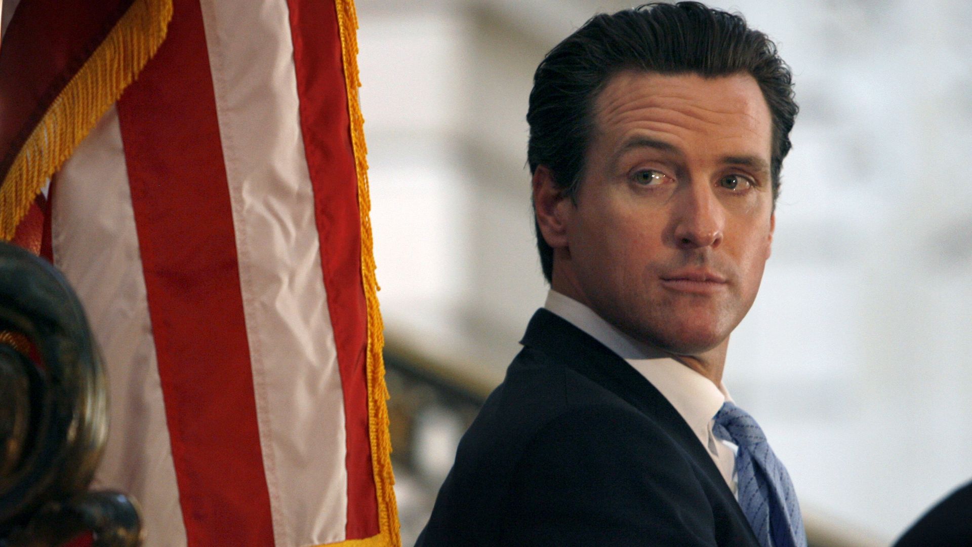 Side profile of a man in a dark suit and light blue tie, looking to the right, with a red-and-white striped flag with gold fringe in the foreground.
