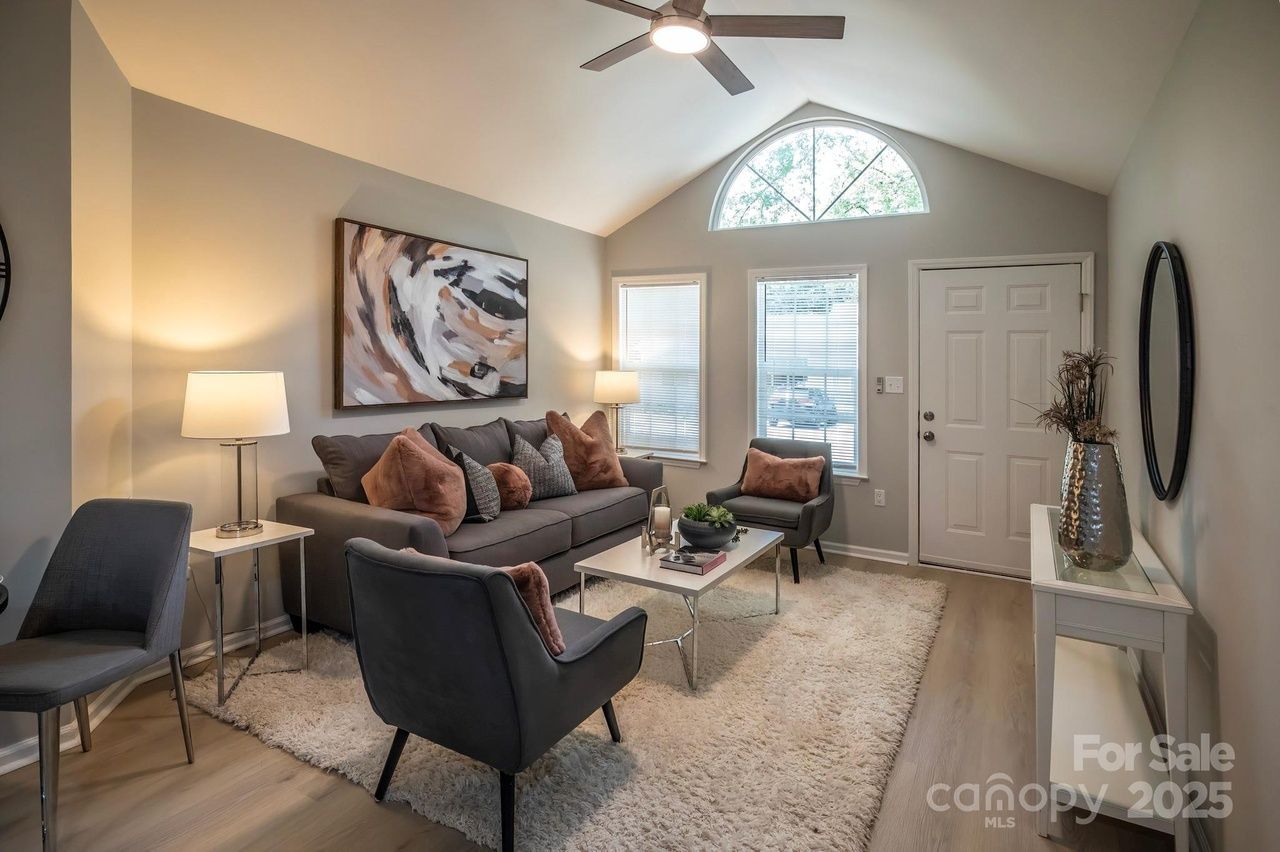 Cozy living room with gray couch, two gray chairs with pink cushions, a white coffee table, two windows, beige walls, abstract wall art, ceiling fan, and a white shag rug.