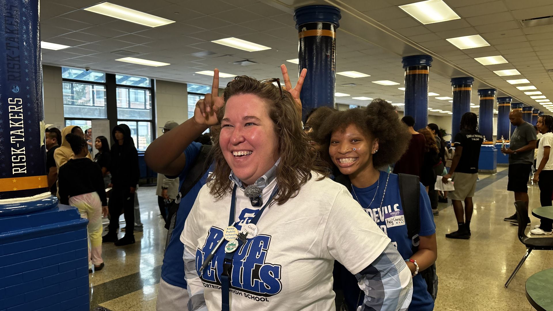 A smiling woman wearing a white Blue Devils shirt stands indoors with a young woman behind her giving bunny ears. People are visible in a school hallway with blue pillars and a microphone with 13 WTHR logo.