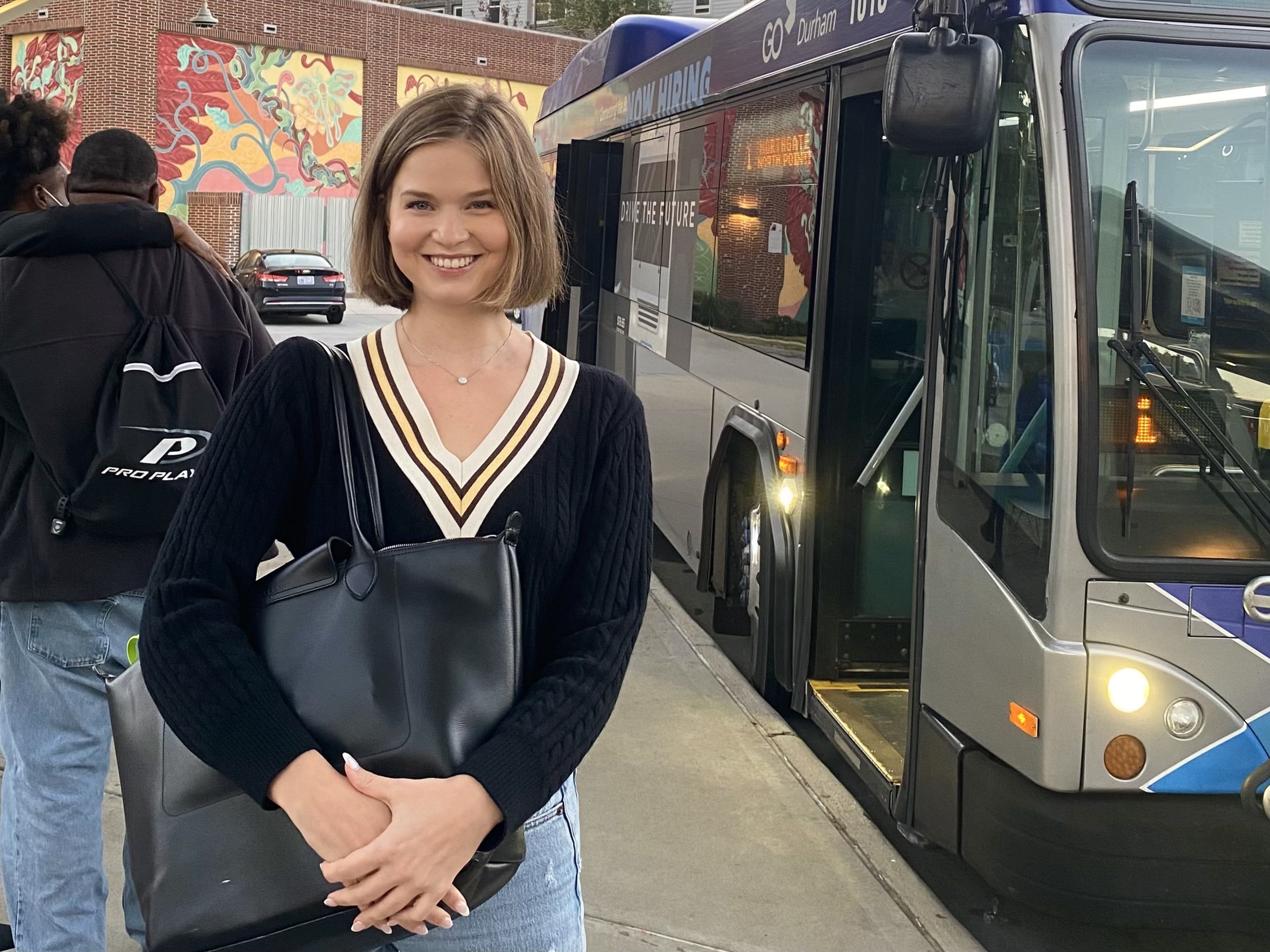 Smiling woman with short blond hair in a navy V-neck sweater, holding a black bag, stands at a bus stop beside a modern bus. Colorful mural in background.