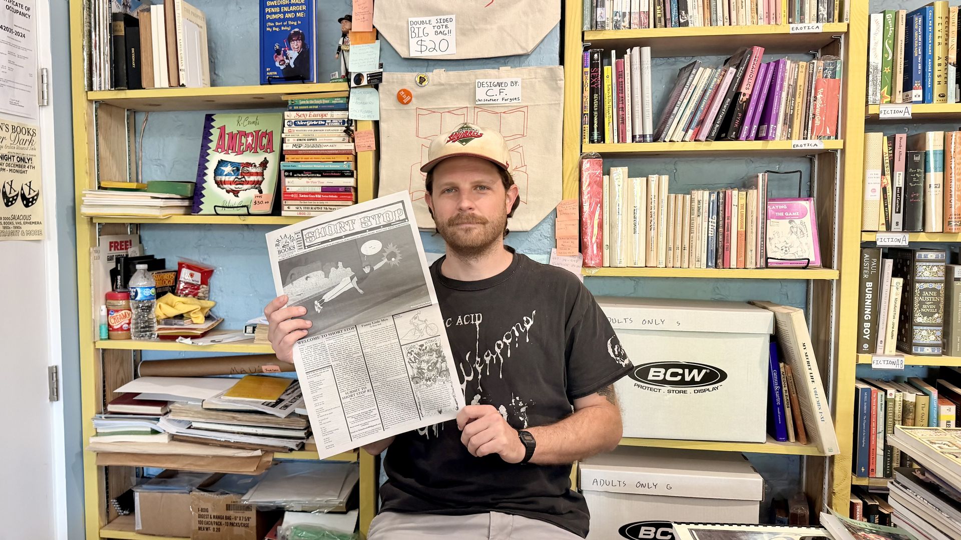 A man with a beige cap and a beard sits in a colorful bookstore, holding a black-and-white newspaper titled "Shorty Stop." Shelves filled with books, magazines, and boxes surround him.