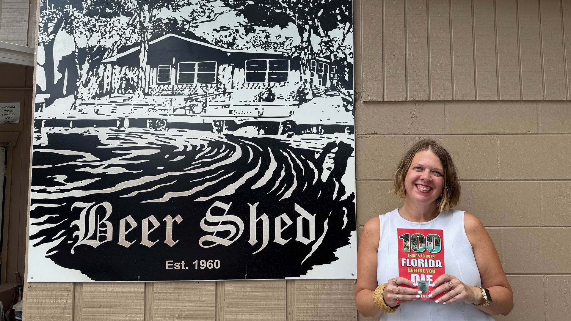 Smiling woman in a white sleeveless top stands beside a large black-and-white mural of a house and trees on a beige wall; mural shows Est. 1960. She holds a red brochure about Florida.