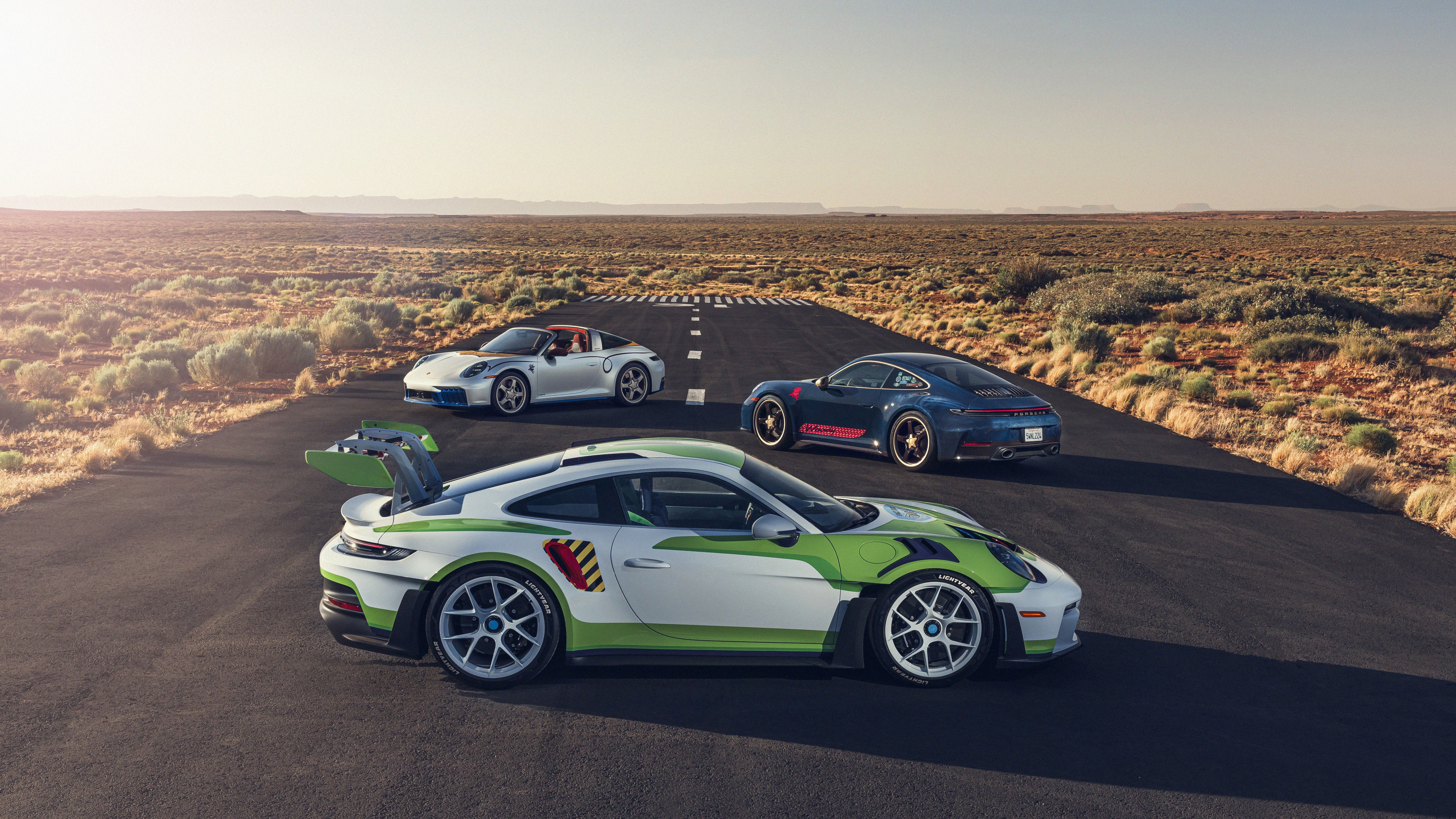 Wide desert highway with several Porch sports cars parked on the asphalt; white convertible, blue Porsche, and lime-green/white race car in the foreground, with scrub brush and mesas on horizon.