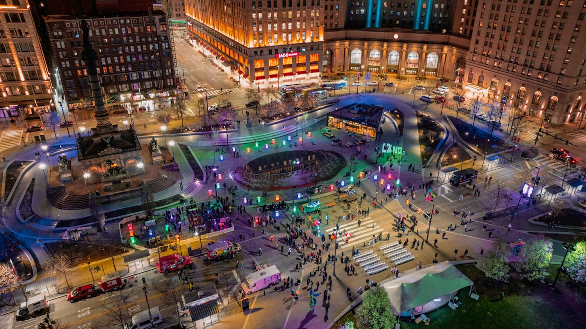 Aerial view of a lively nighttime city square with colorful illuminated columns, a central fountain, a monument, and crowds of people around food trucks and seating areas under surrounding lit buildings.