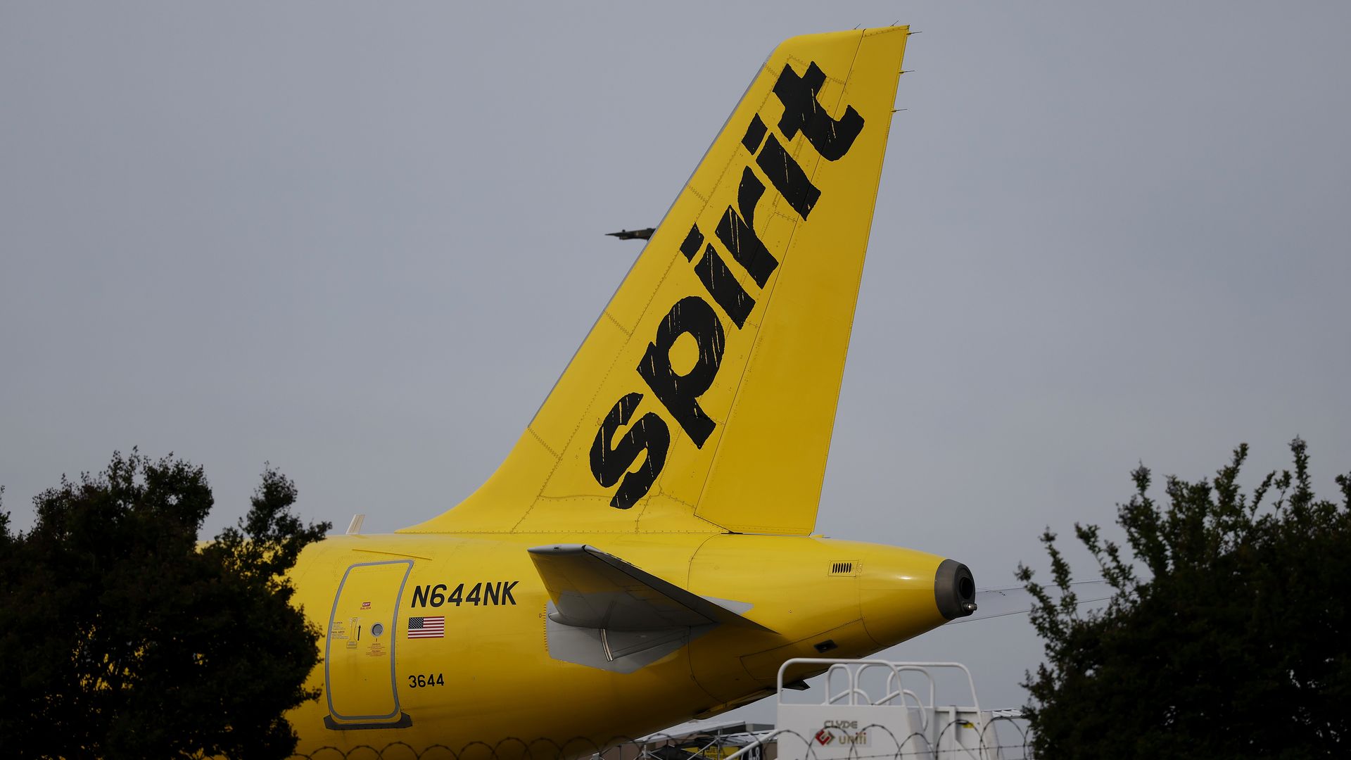 Close-up of a bright yellow airplane tail with a black GOL logo, registration N644NK, at an airport. A fence and trees frame the foreground, with a small aircraft visible in the sky.