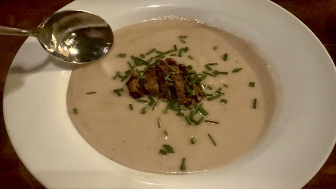 Creamy soup in a white bowl garnished with chopped green herbs and small brown pieces, with a shiny spoon resting on the bowl edge on a wooden table.