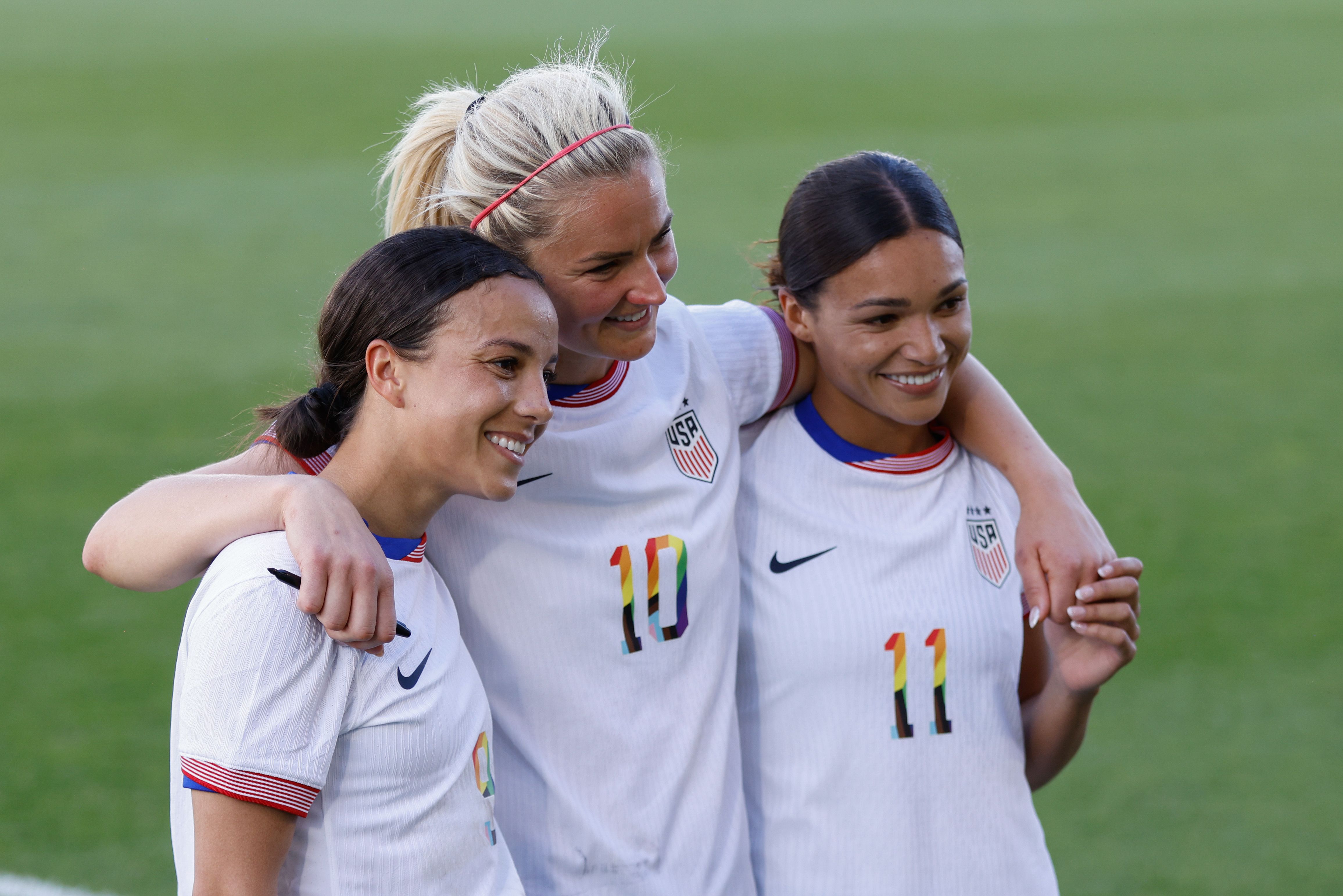 Three women in white jerseys smile while posing for a photograph while standing on a field. 