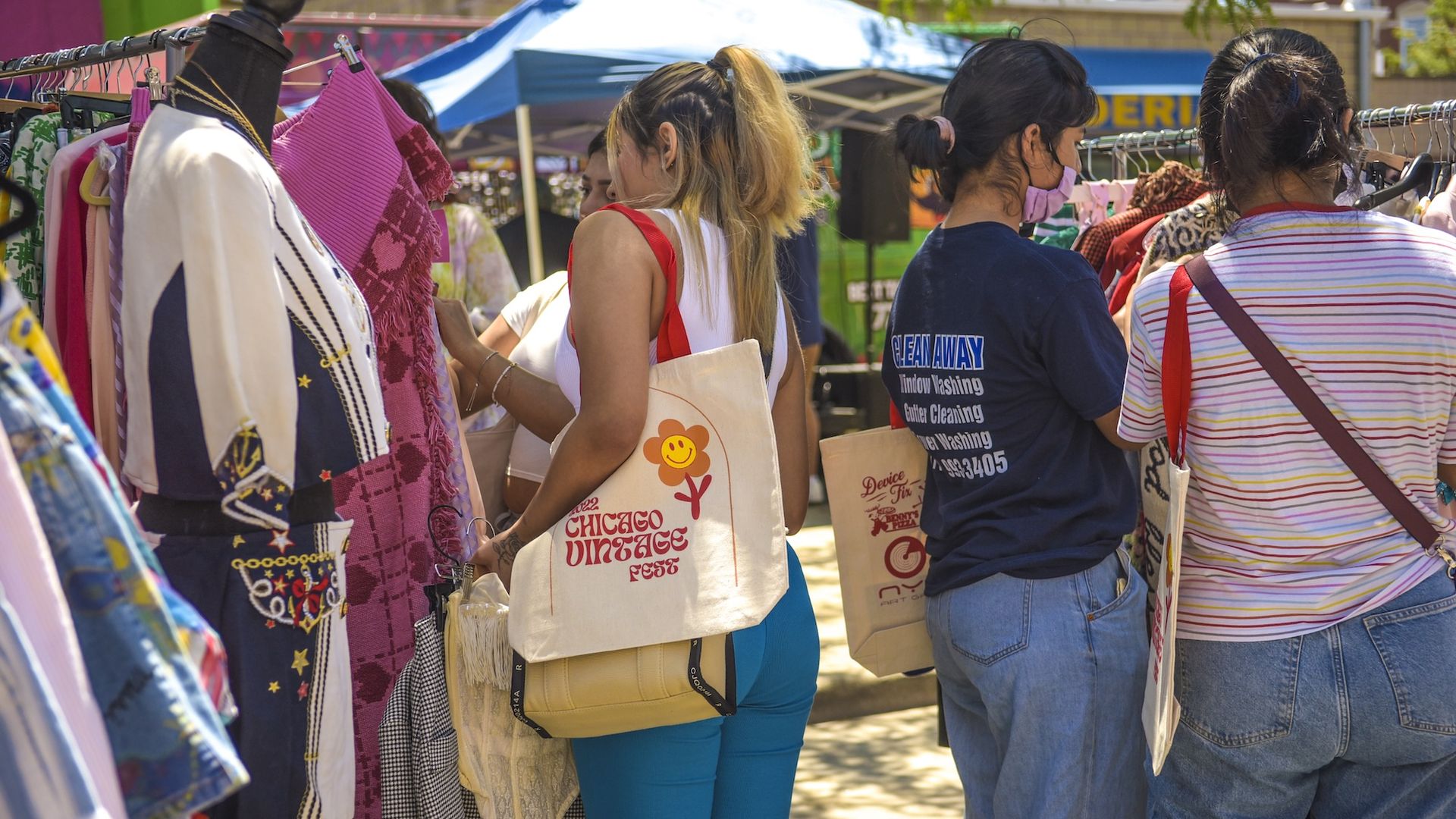 People browsing vintage clothes at an outdoor market with colorful garments on racks and tote bags, including one that reads "Chicago Vintage Fest." Sunlit scene with tents in background.