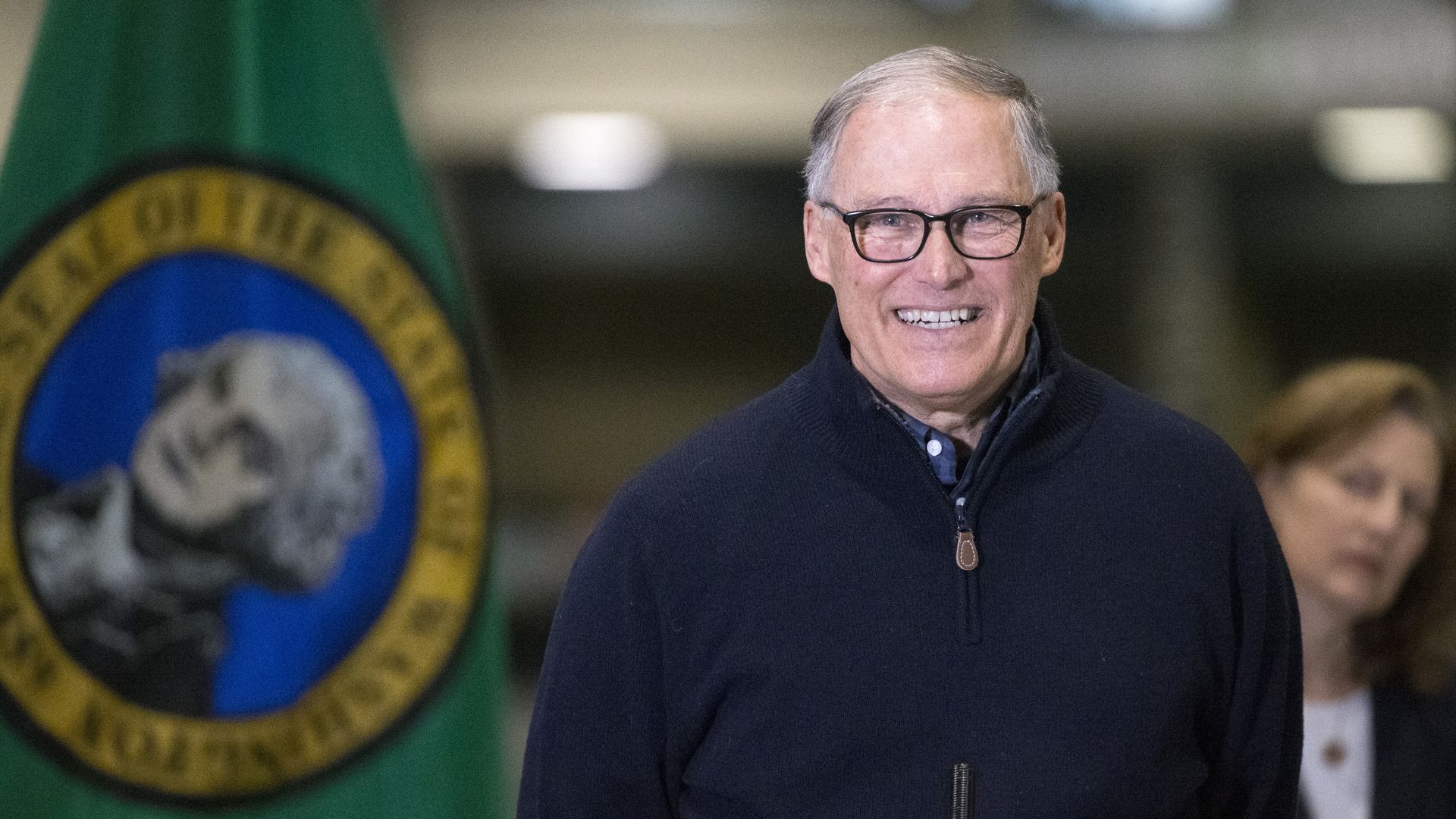 Gov. Inslee smiles while wearing a pullover while next to a flag of the state of Washington.