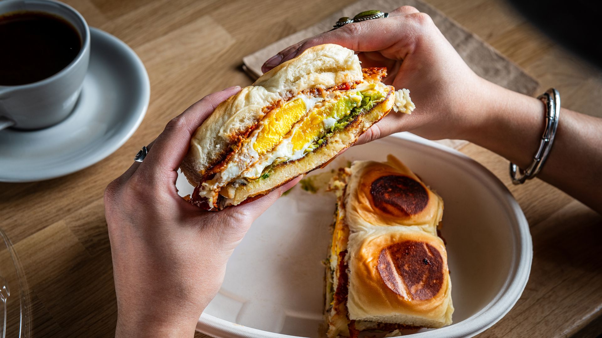 A person with rings and bracelets holds a toasted breakfast sandwich filled with egg, cheese, and avocado, at a wooden table with a cup of coffee and pastries on a plate nearby.