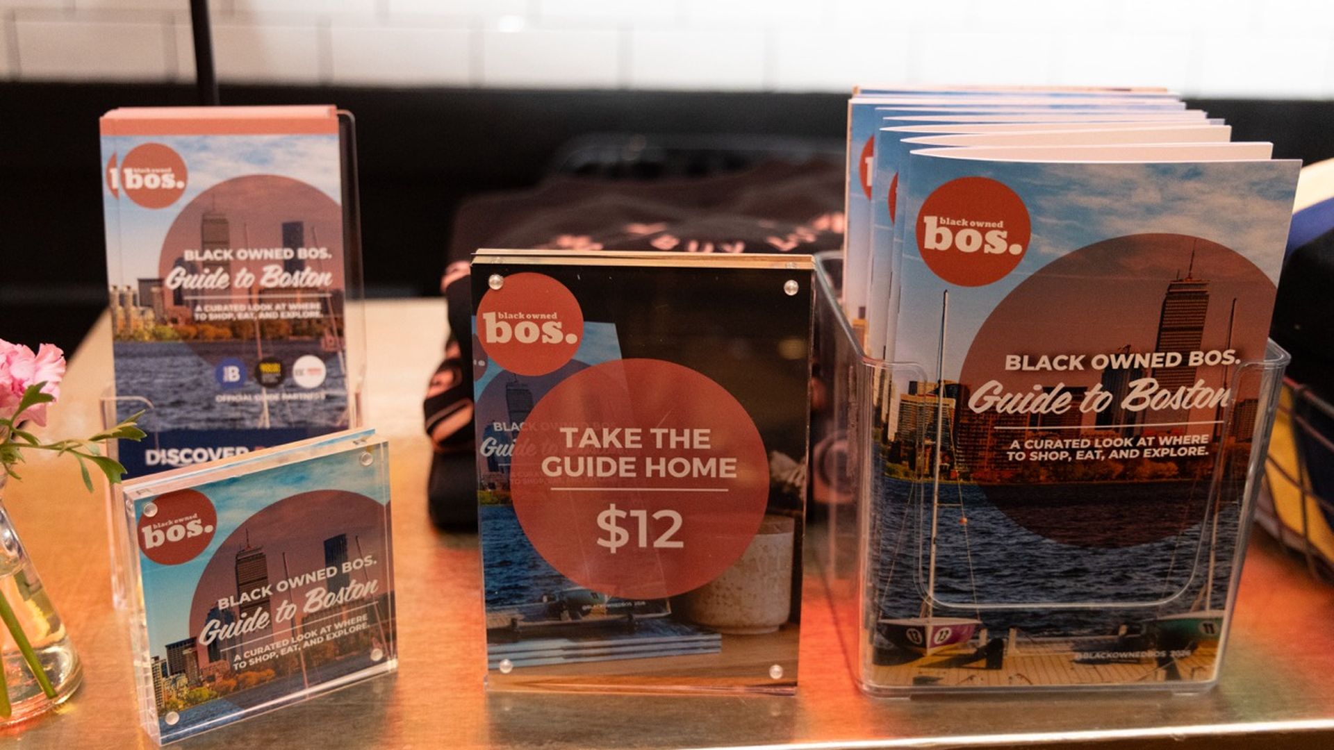 Display of "Black Owned BOS. Guide to Boston" brochures on a shelf, featuring blue cityscapes and red circular logos. A central glass plaque reads "Take the Guide Home" for $12.