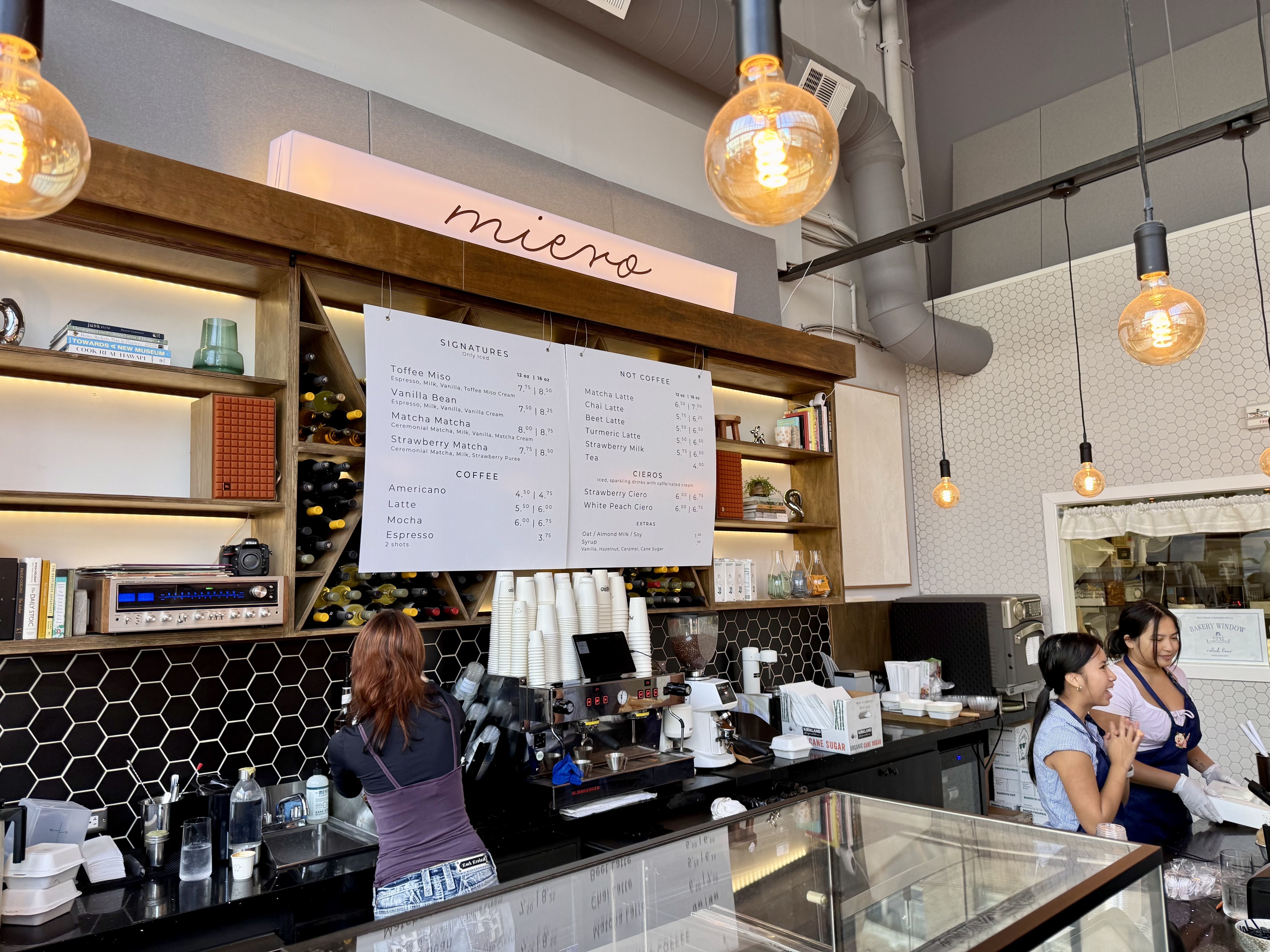 Cafe counter with hexagonal black backsplash, warm hanging bulbs, a cursive sign reading "mi amore", menu boards, and staff serving customers.