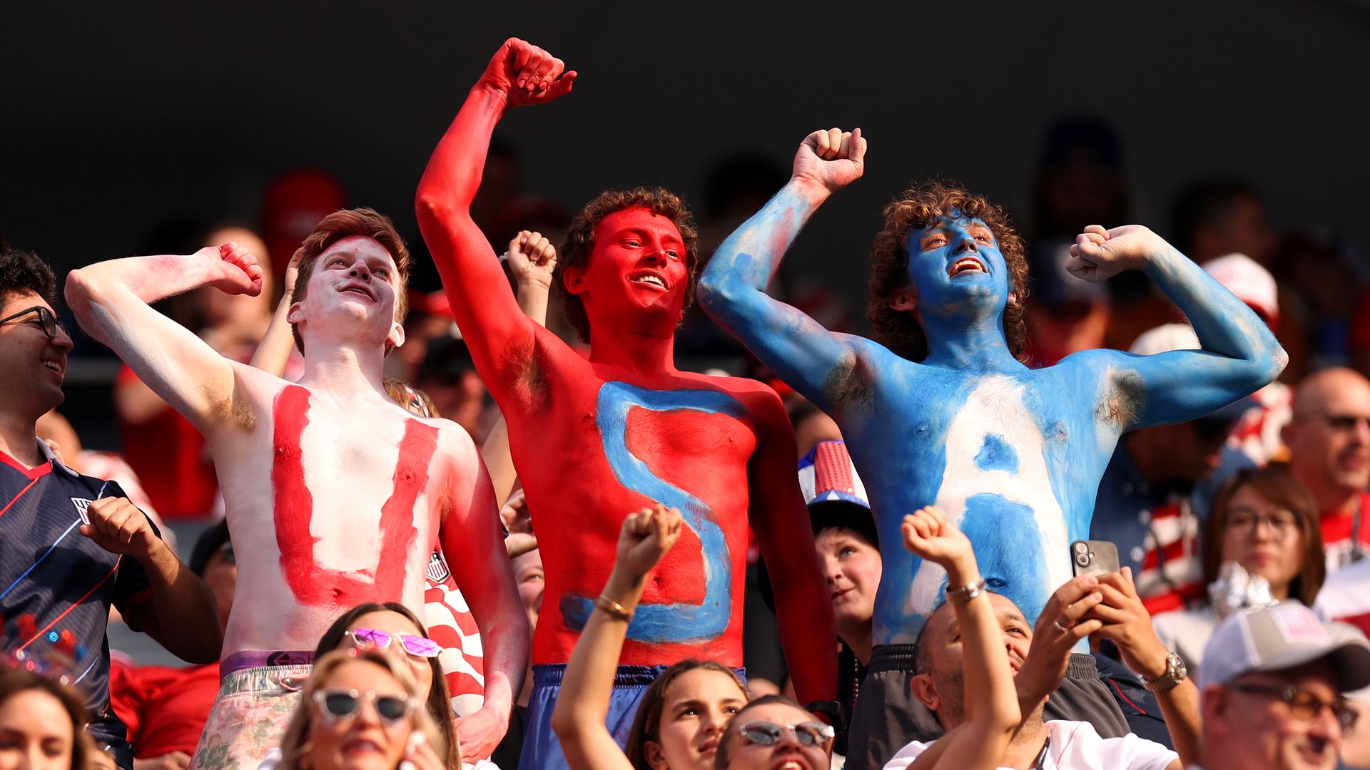 Three men cheering at a game painted in Red white and blue with the letters U S and A painted on their chests.