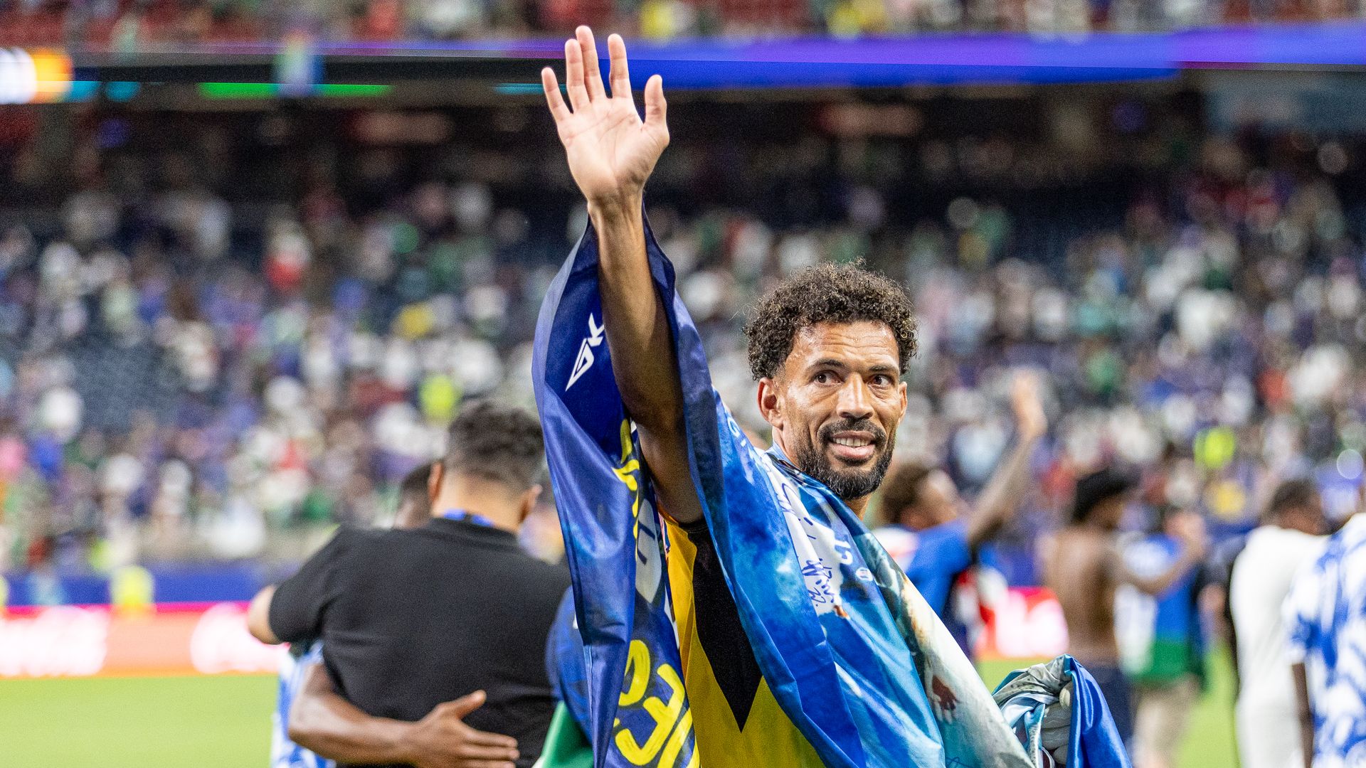 An athlete raises his hand while crowds cheer in a stadium; a blue and yellow flag drapes his shoulders as he celebrates.