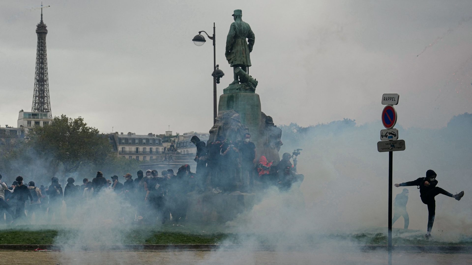 Wide, smoke-filled scene in a Paris square: a bronze statue on a pedestal, crowds at the base, and a tall lattice tower to the left; a street sign reads RAPPEL, with a no-parking sign nearby.