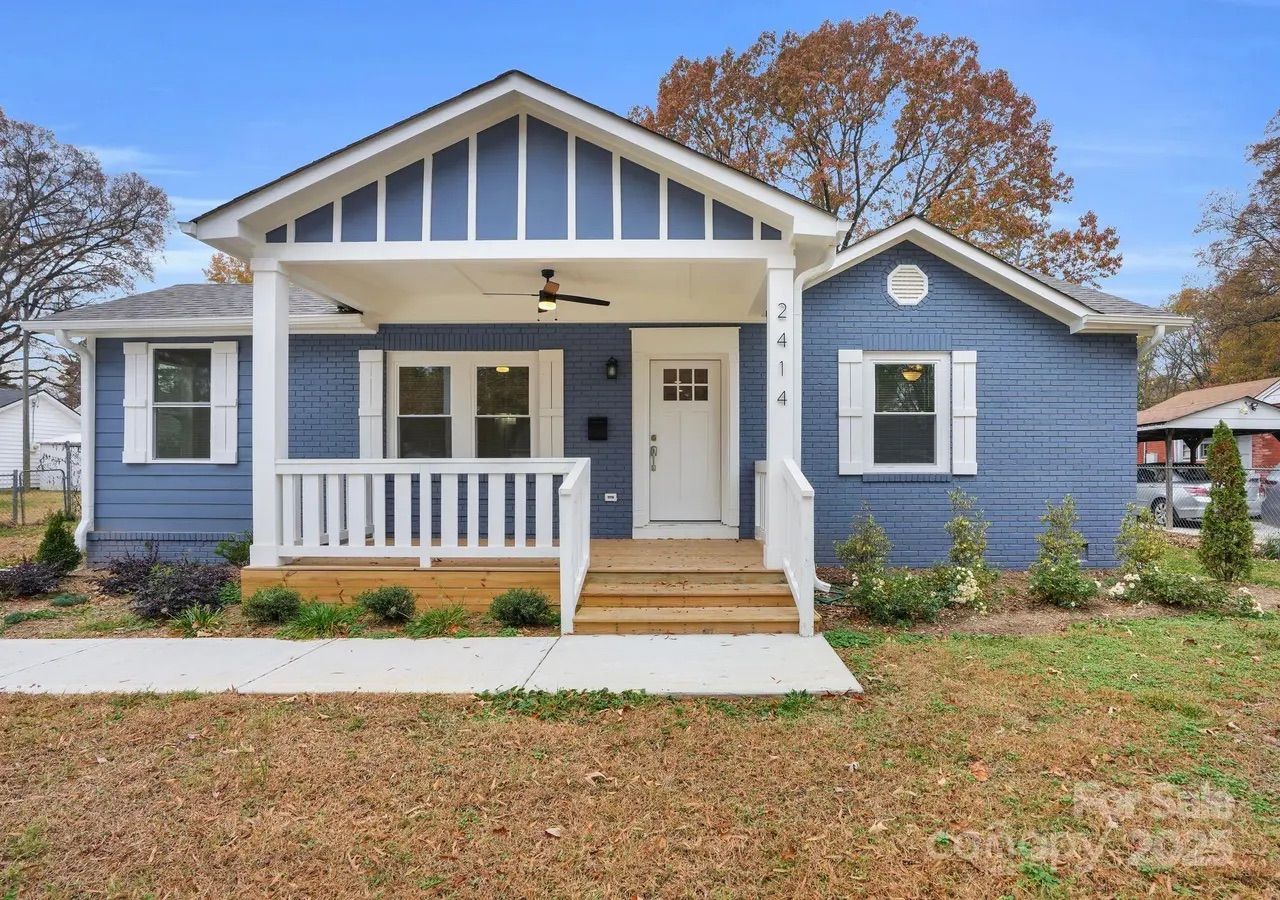 Blue brick house with white trim, front porch with white railings, wooden steps, and a ceiling fan, surrounded by autumn trees and lawn.