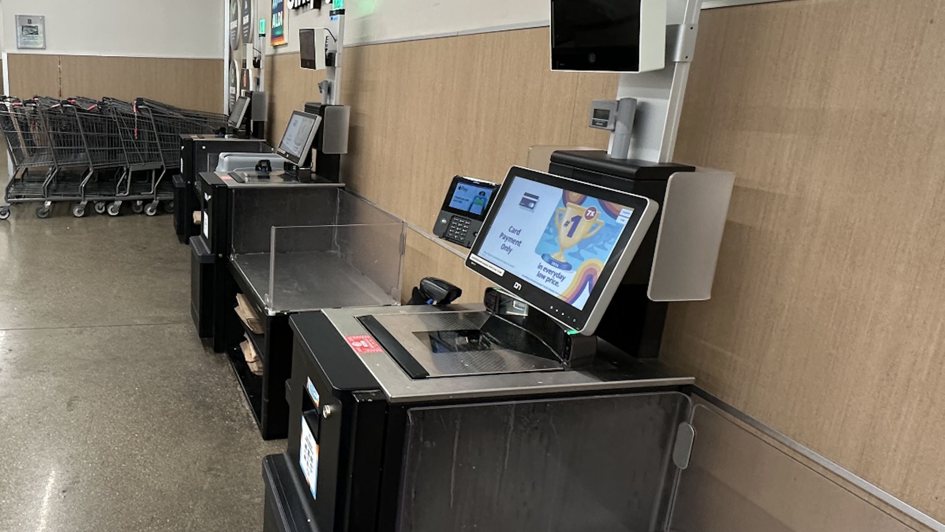 Row of black self-checkout machines with card payment screens in a store, empty shopping carts lined up in the background, beige wall behind the machines.