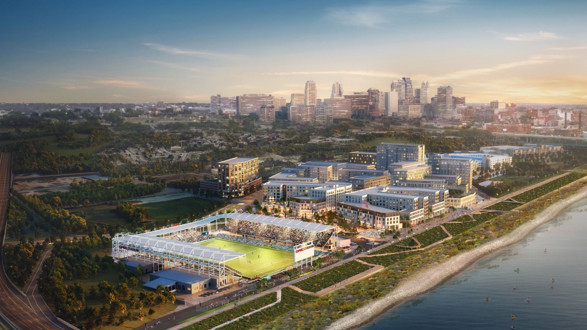 Aerial view of a lit soccer stadium by a river at sunset, surrounded by modern buildings and green spaces, with a city skyline in the distance under a partly cloudy sky.