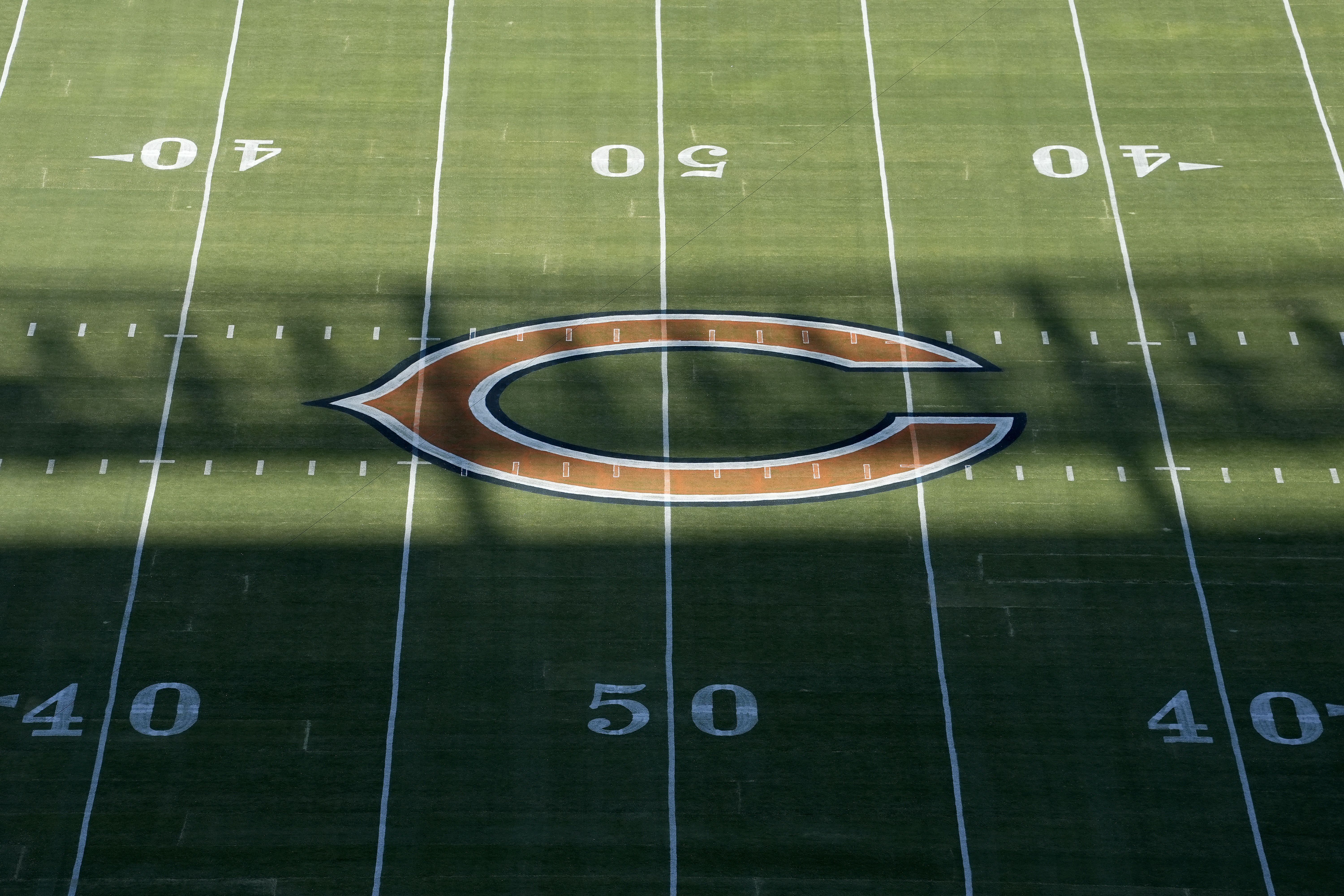 Aerial view of a green football field showing the 40, 50 yard lines and a large orange and navy blue 'C' logo at the center, partially shaded by a shadow.