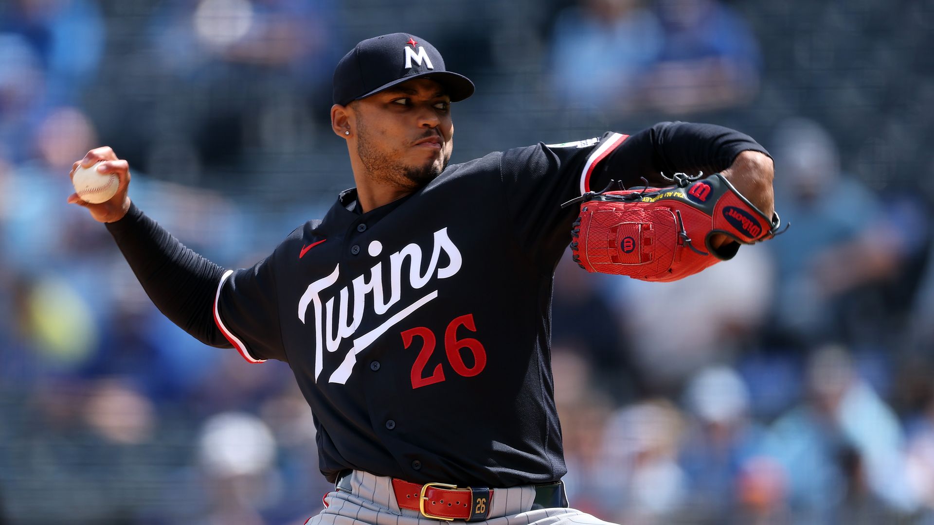 Baseball pitcher from the Minnesota Twins in a black uniform with white "Twins" script and red number 26, mid-pitch with a red glove on the right hand and a ball in the left, crowd blurred behind.
